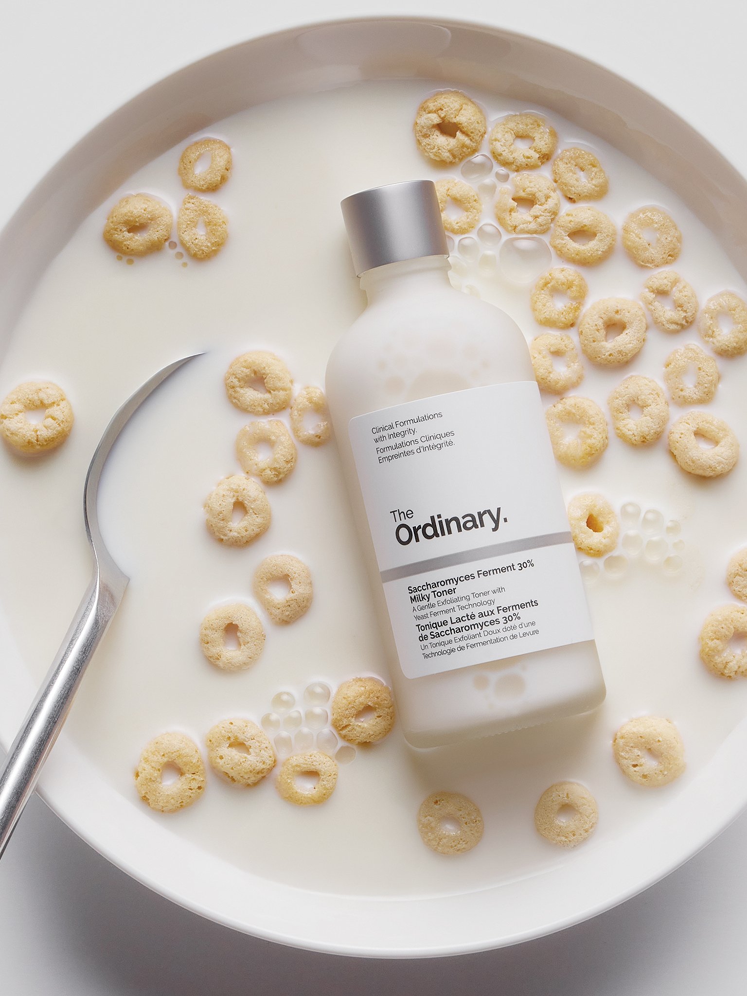 A bottle of The Ordinary facial toner surrounded by floating cereal pieces in a white liquid, on a white surface with a silver spoon nearby.