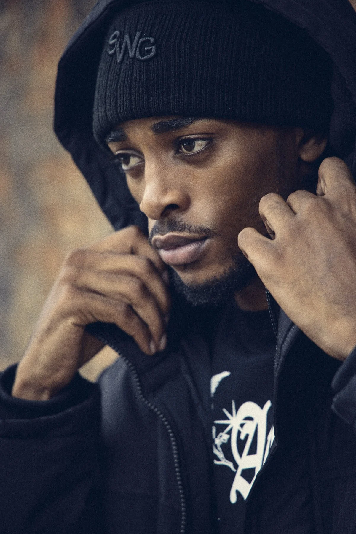 Close-up of a young man wearing a black beanie and hoodie, adjusting the hood while looking down.