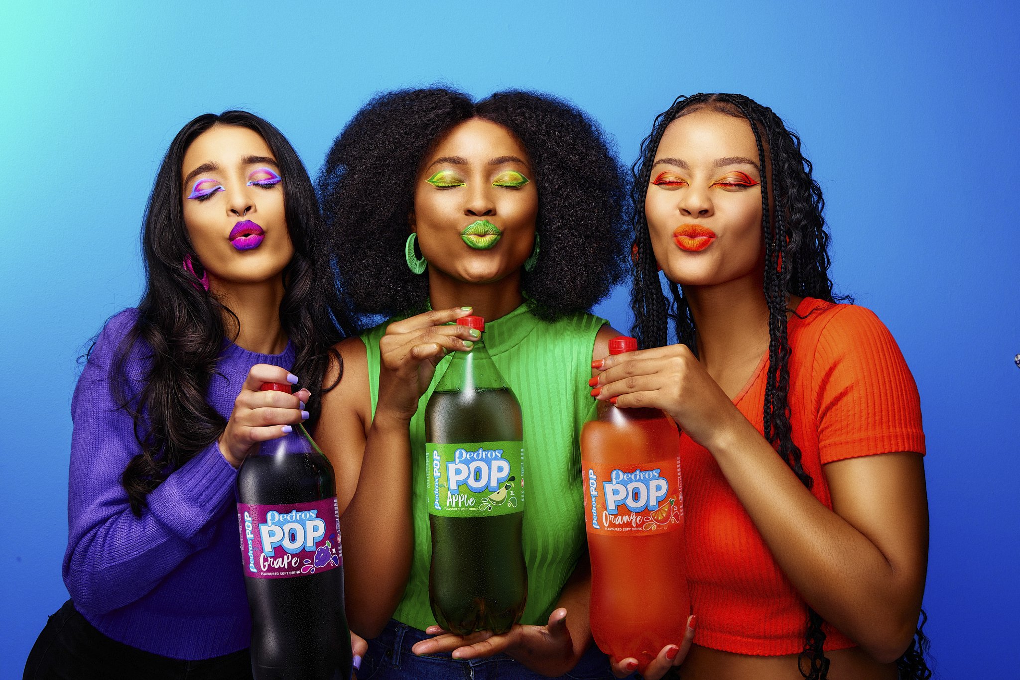Three women with colorful makeup and clothing holding bottles of Pedros Pop flavored soft drinks with a blue background.