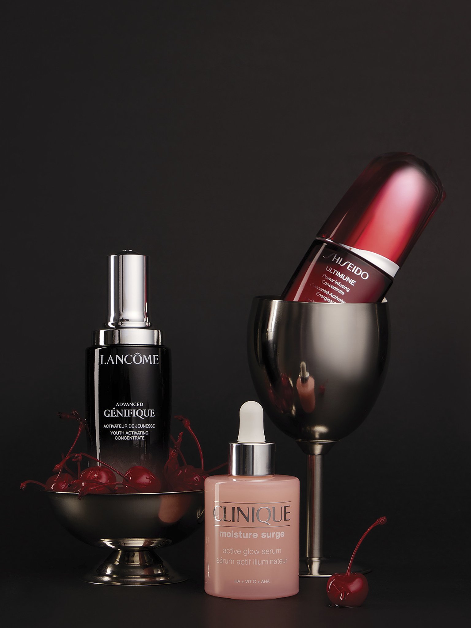 Skincare products, including Lancôme Advanced Génifique Youth Activating Concentrate, Clinique Moisture Surge serum, and Shiseido Ultimune Power Infusing Concentrate, arranged on a black background with cherries and a glass of red liquid.