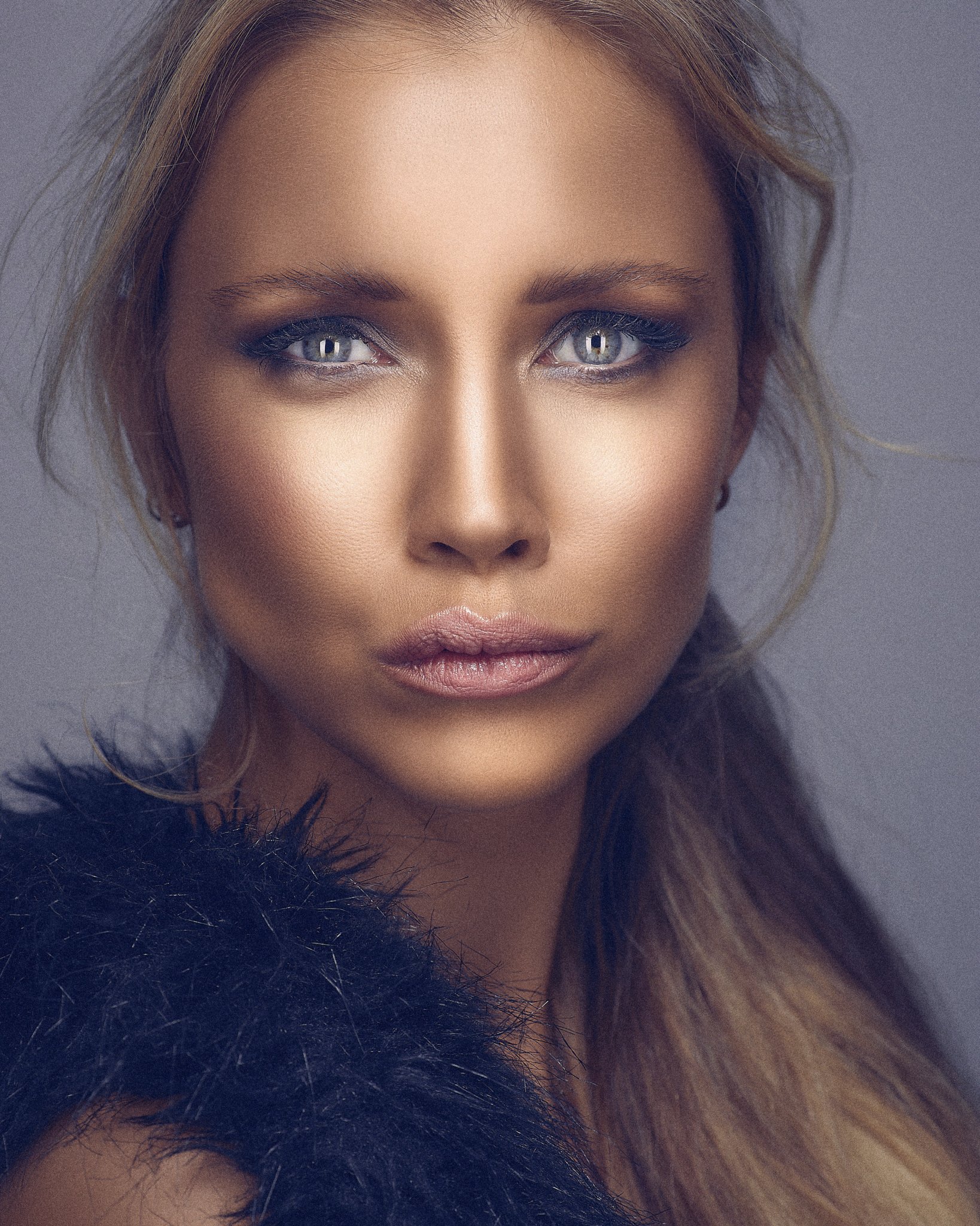 Close-up of a woman with blonde hair and blue eyes, wearing makeup, with a dark fur coat collar visible in the foreground.