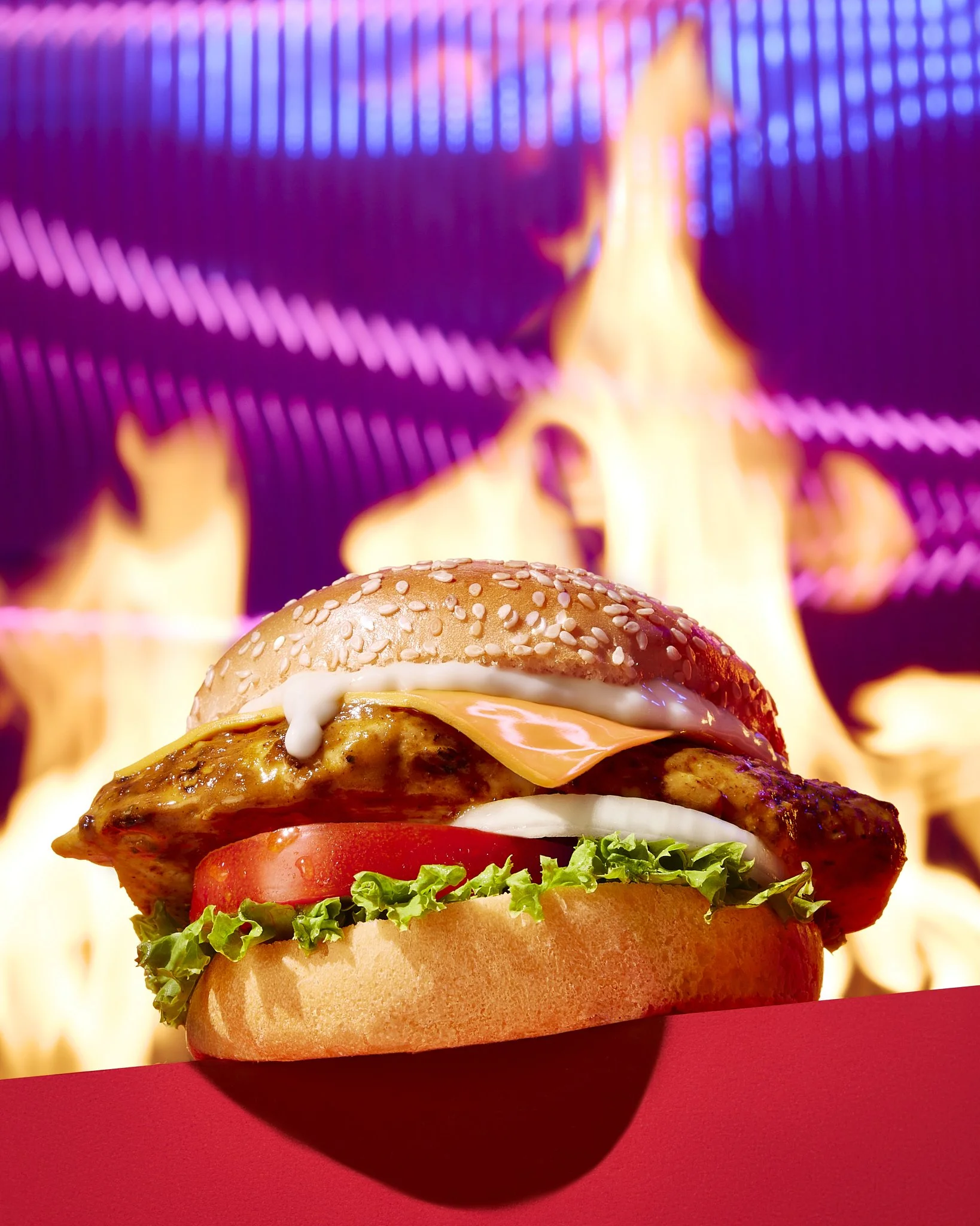 Close-up of a hamburger with sesame seed bun, lettuce, tomato, cheese, and a grilled chicken patty, with flames in the background.