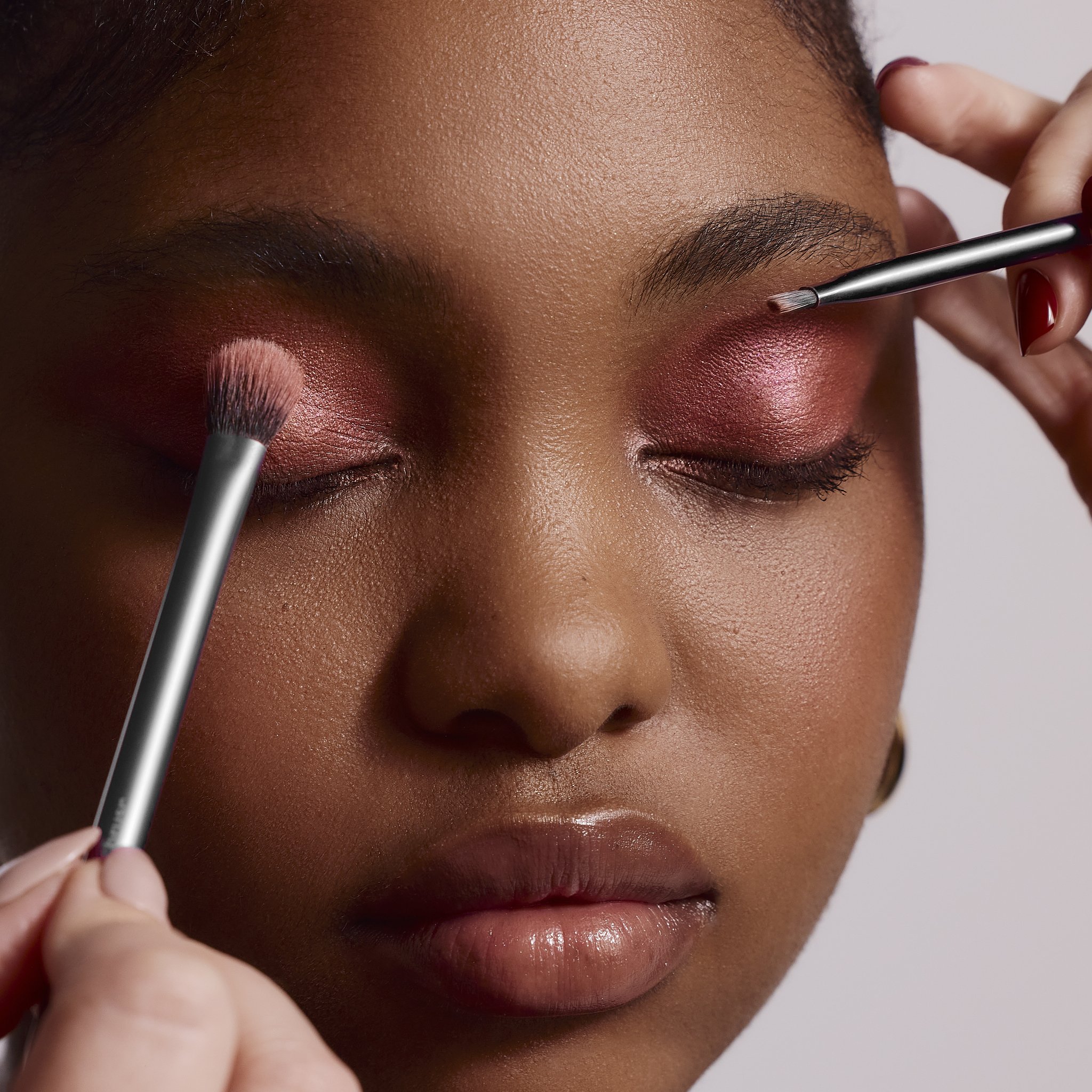 Close-up of a person with dark skin getting their eyebrows groomed with a small brush by a person's hand. The person has natural eyebrows, closed eyes, and wearing makeup with pink eyeshadow.