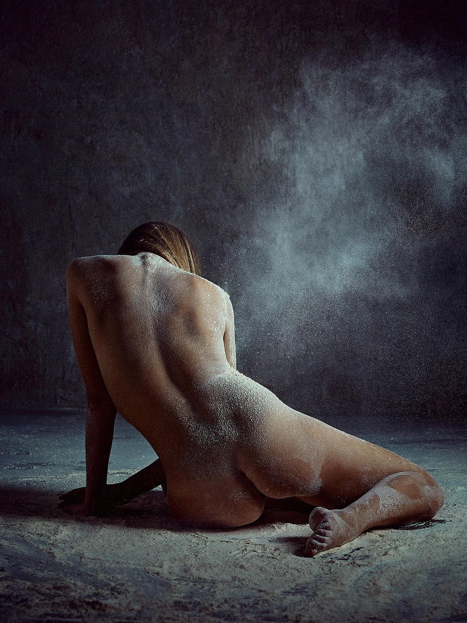 A nude person crouches on the sand, with their back to the camera and their head bowed, surrounded by a misty atmosphere.
