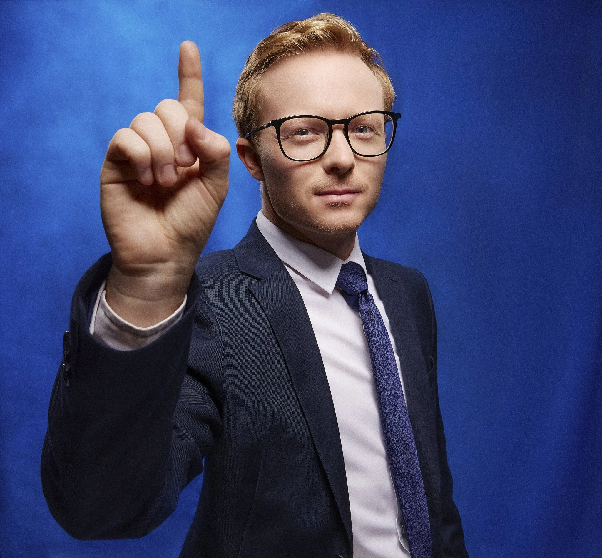 Democratic Alliance Chris Pappas with blond hair, wearing glasses, a dark suit, white shirt, and purple tie, is raising his right hand with his index finger pointing upwards against a blue background.