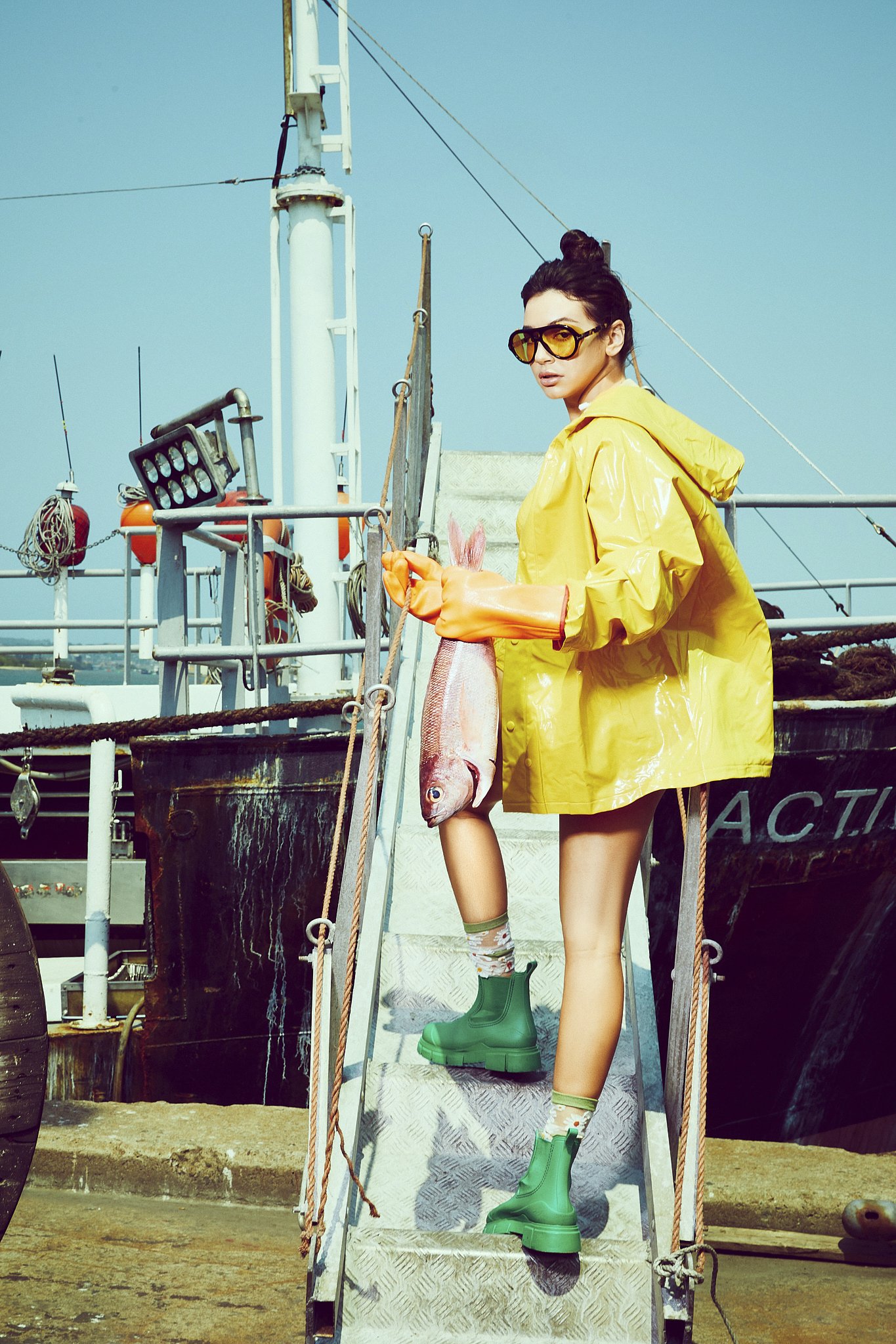 Woman wearing yellow raincoat, green rain boots, and sunglasses holding a fish while standing on a metal ladder on a boat.
