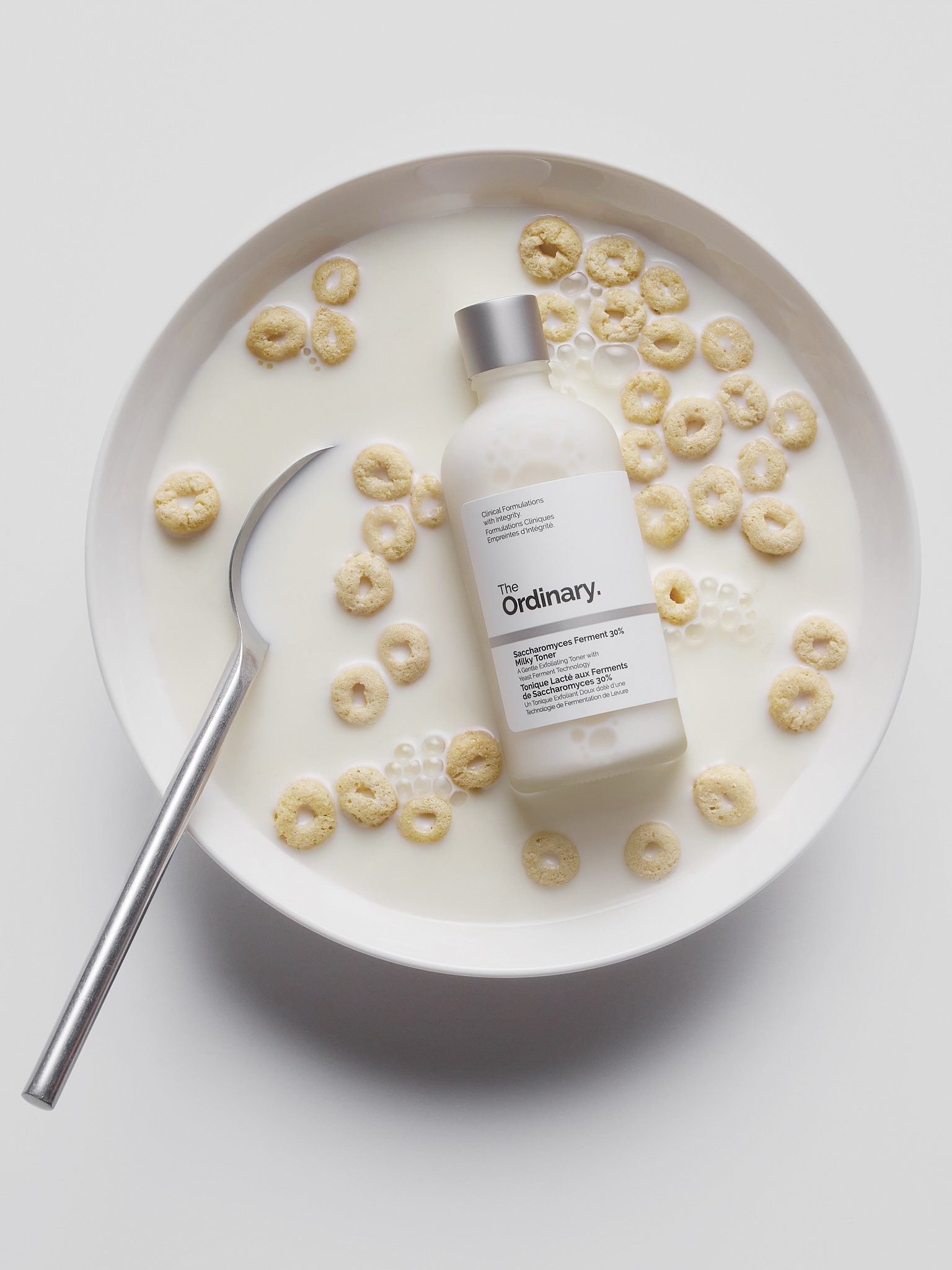 A white bowl filled with milk and cereal, with a metal spoon, a bottle labeled 'The Ordinary' skincare product, and scattered cereal pieces.