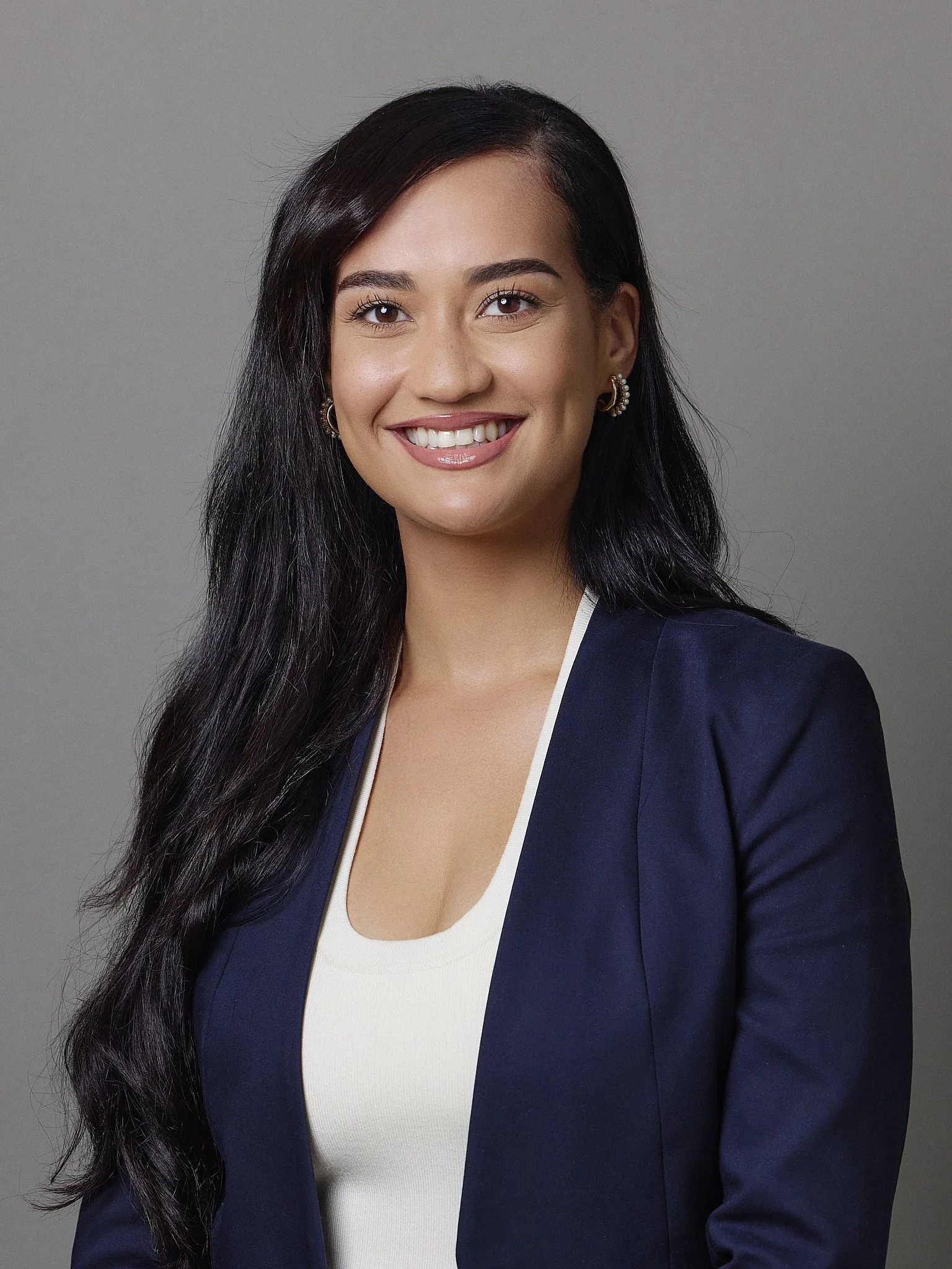 Portrait of a smiling woman with long black hair, wearing a navy blazer and a white top, against a plain gray background.