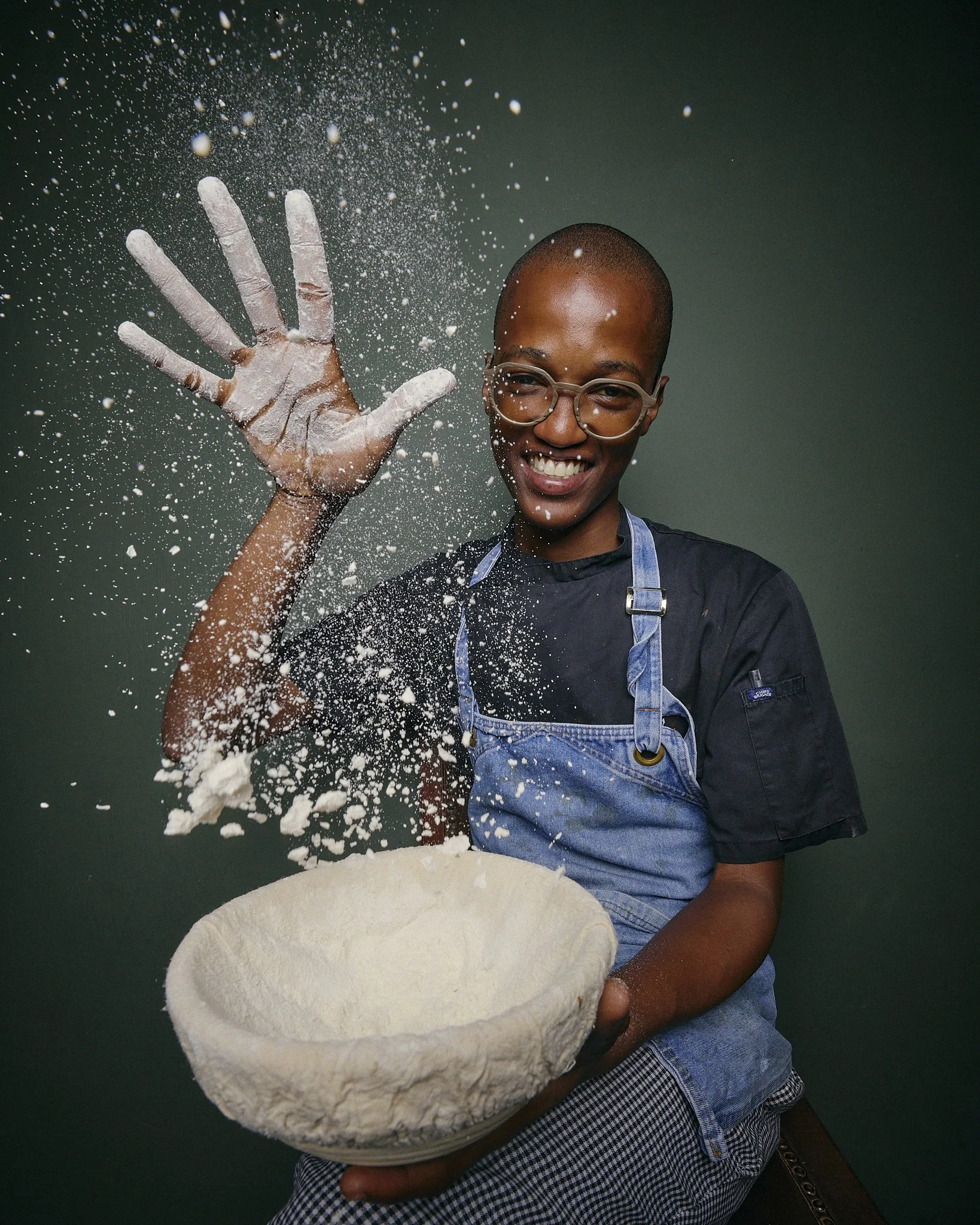 A person with glasses, smiling and wearing a chef's apron, is holding a large bowl of dough while tossing flour into the air against a dark background.