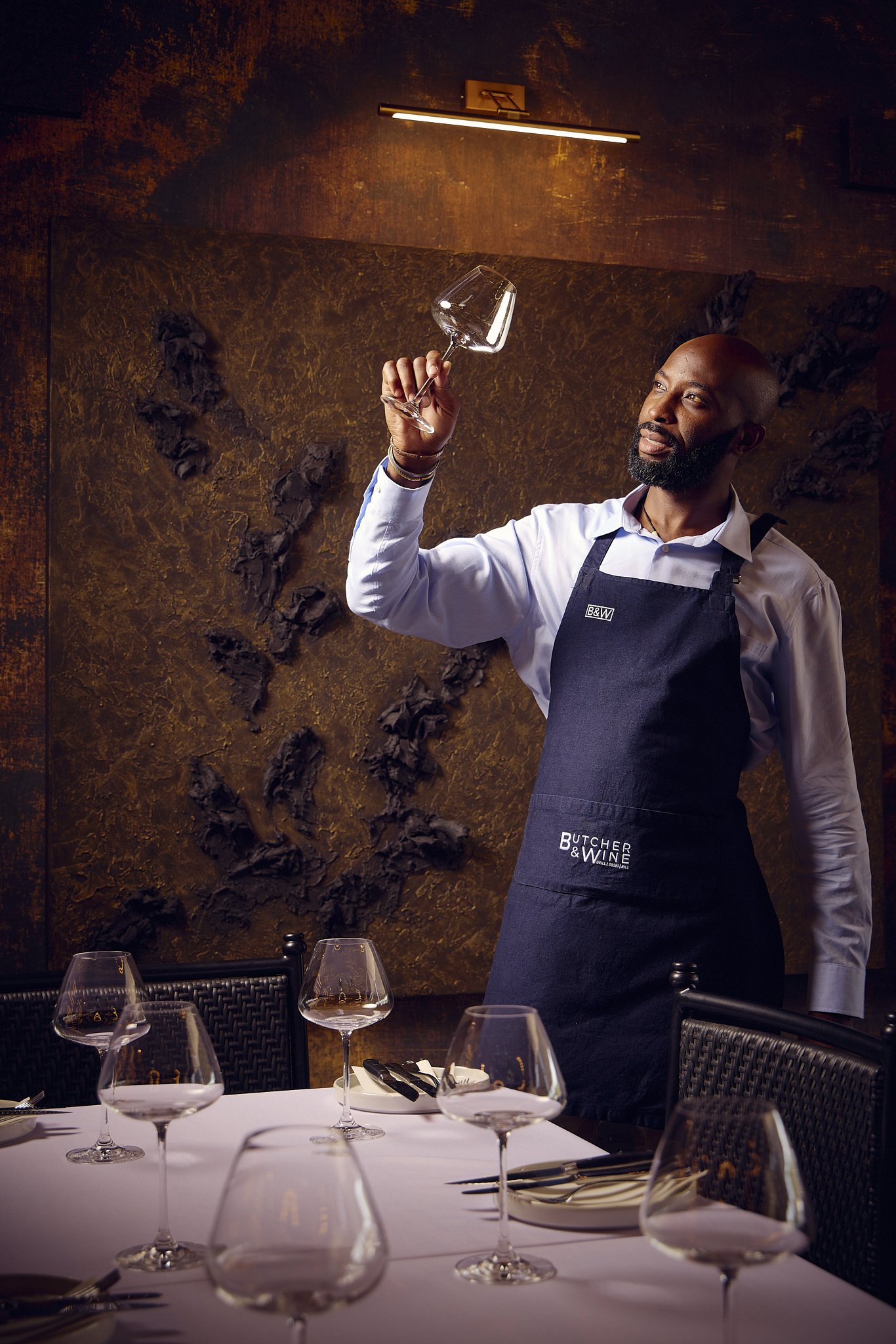 A man in a white shirt and black apron, standing at a restaurant table, holding a wine glass. The table is set with multiple wine glasses, plates, and utensils. The background features a textured, brownish wall.