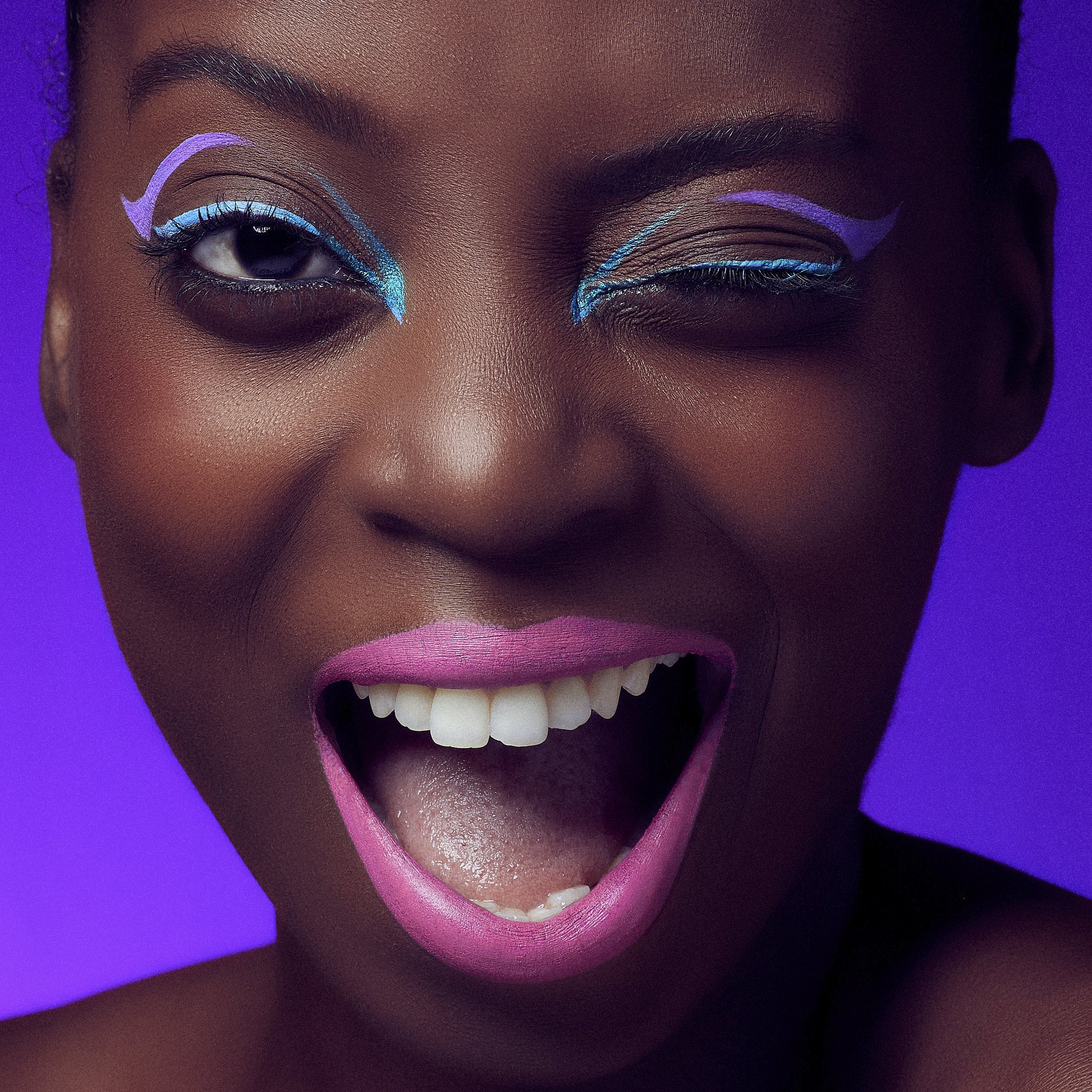 Close-up of a person's face with bright neon-style makeup, including blue and purple eyeliner and pink lipstick, against a purple background.