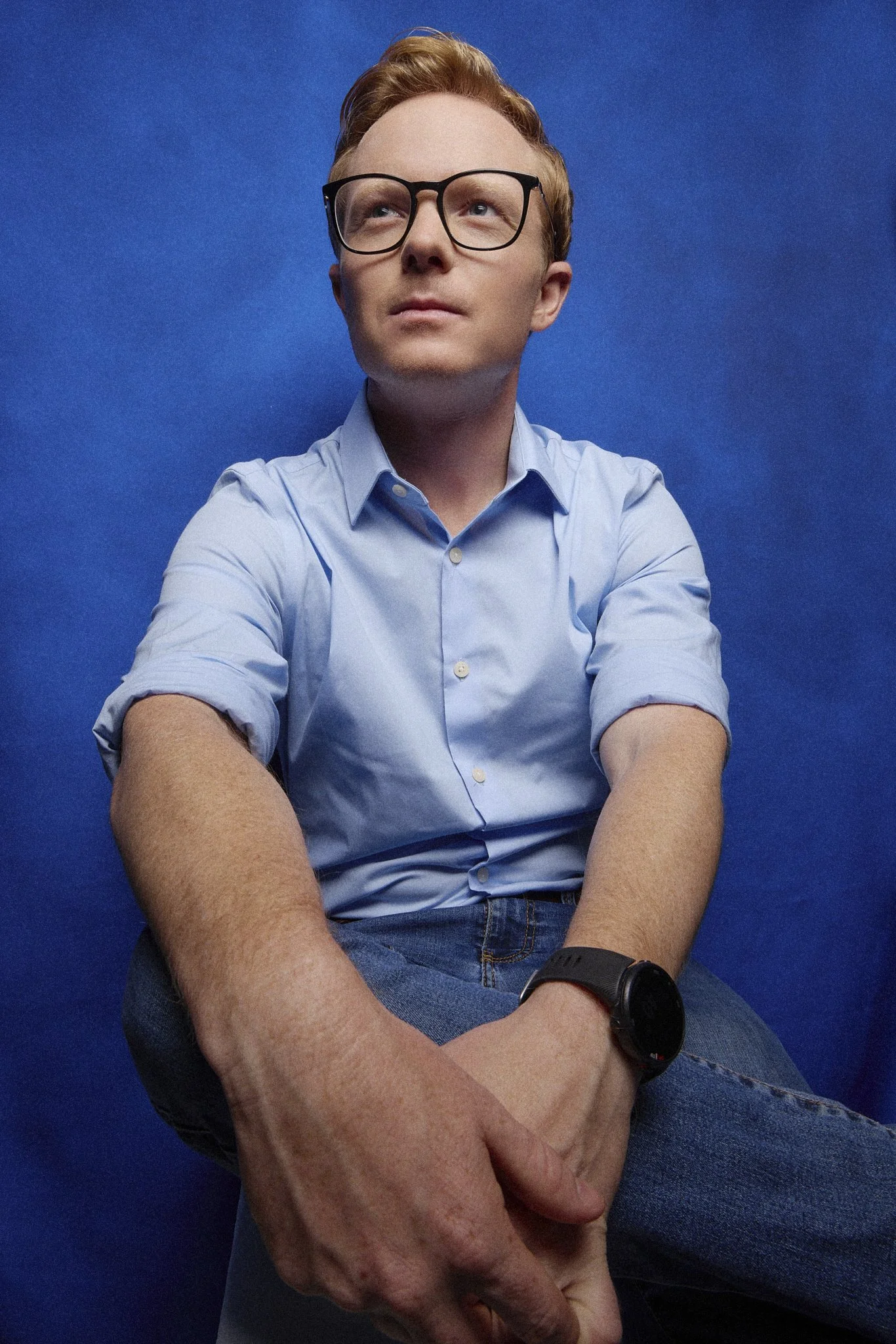 Democratic Alliance Chris Pappas with red hair, glasses, wearing a light blue button-up shirt and jeans, enjoys a moment of reflection against a solid blue background.