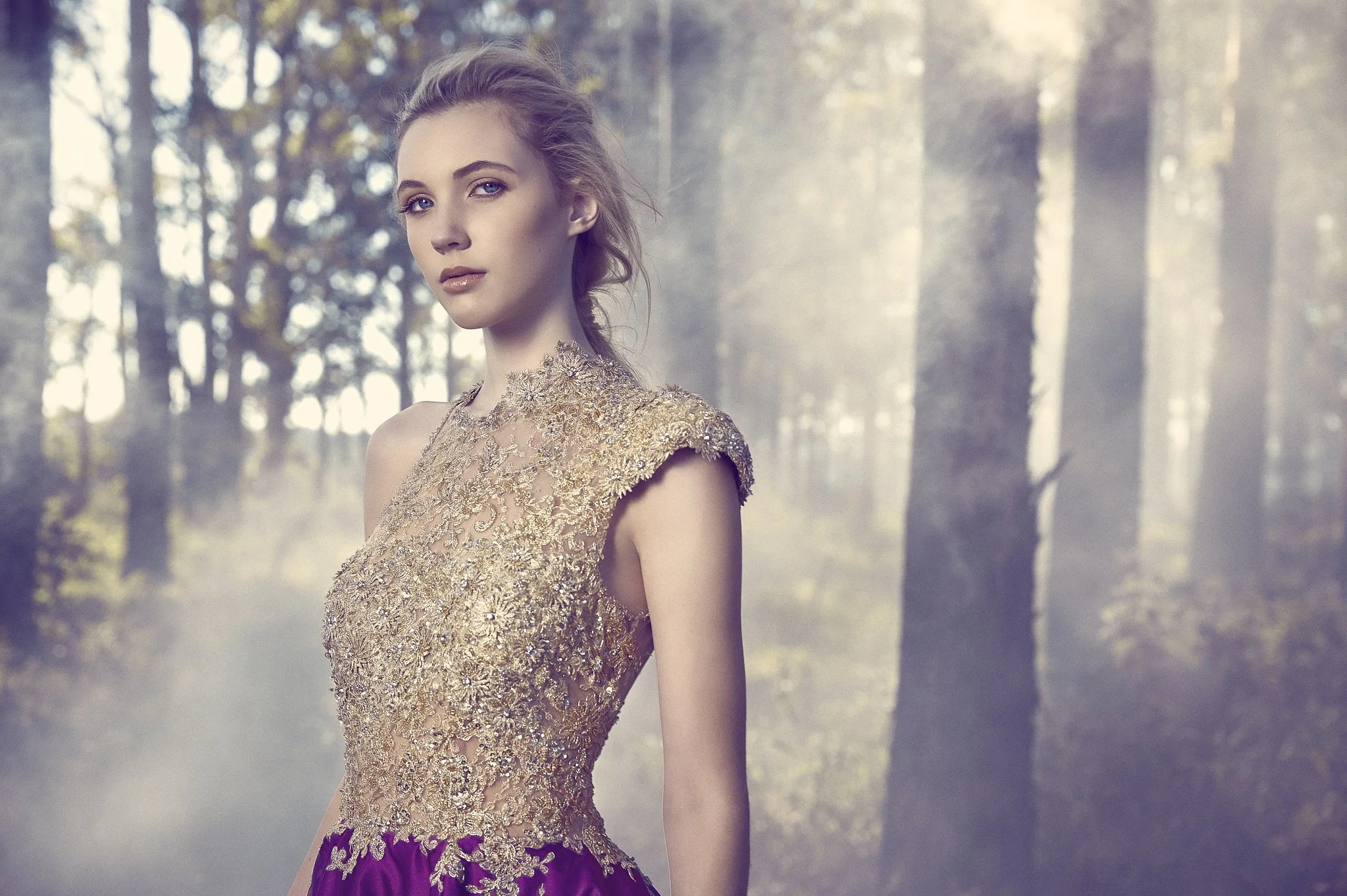 A young woman with blonde hair, blue eyes, and light makeup, wearing a gold floral lace dress, standing in a misty forest with tall trees in the background.