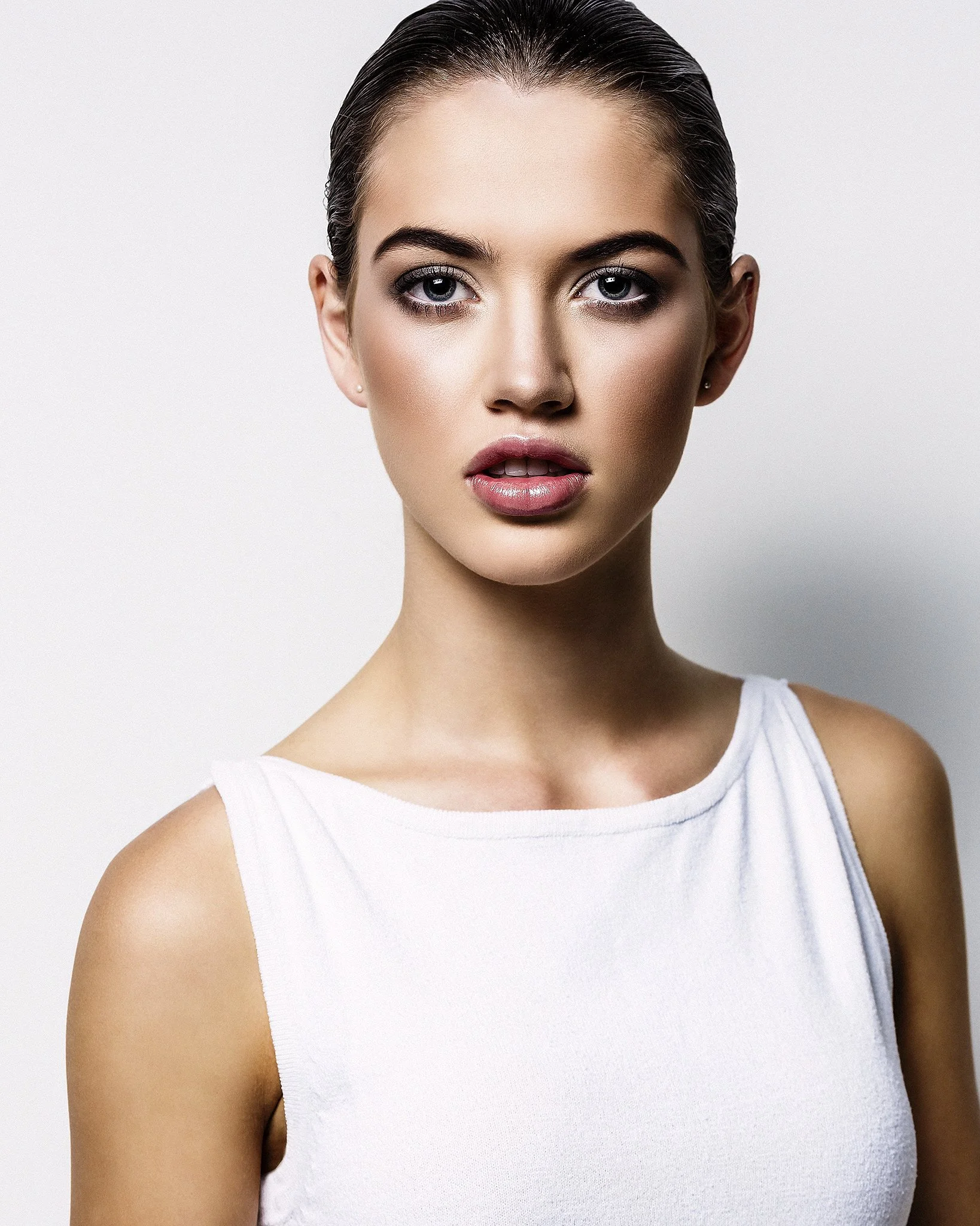 A young woman with slicked-back dark hair and striking blue eyes is wearing a simple white tank top, standing against a plain gray background.