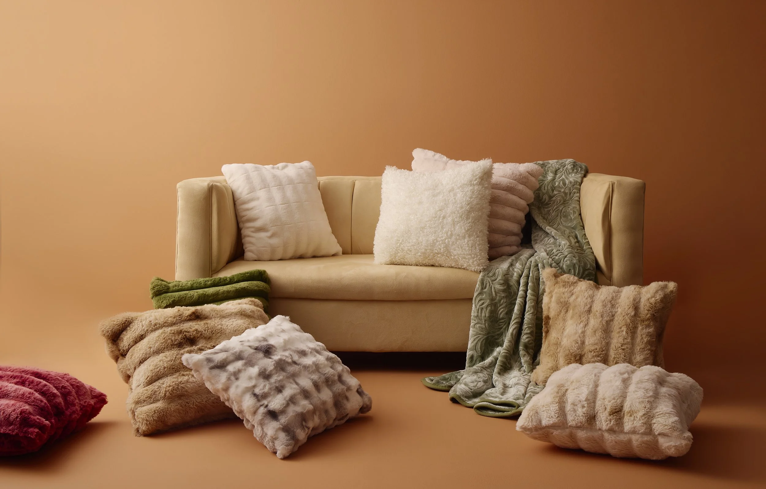 Sheet Street advert showing a beige sofa with various fluffy and textured pillows against a peach-colored wall, surrounded by soft faux fur and plush blankets in different colors on the floor.