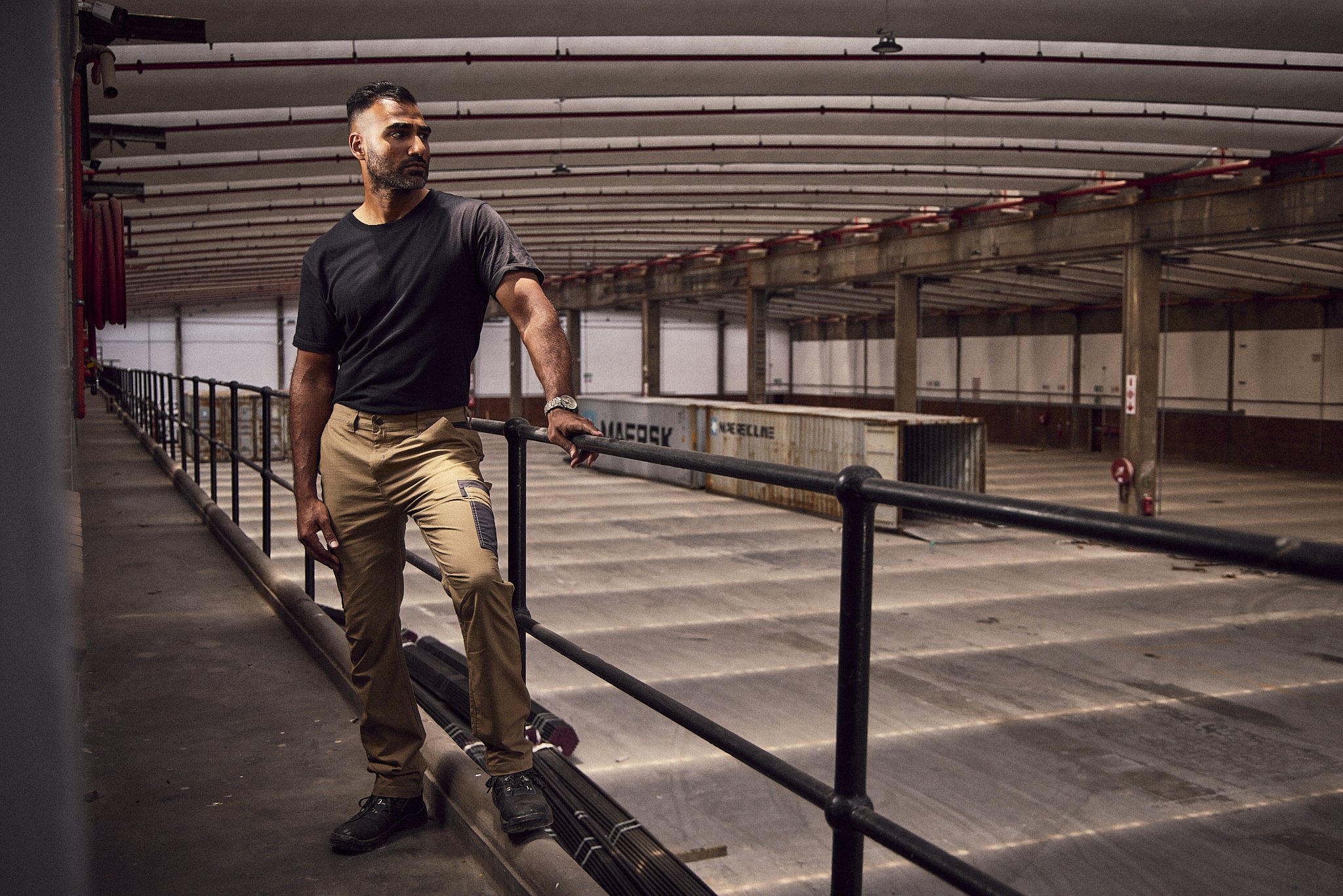 A man in a black t-shirt and beige pants leaning on a safety railing in an underground parking garage, with concrete pillars and cargo containers visible in the background.