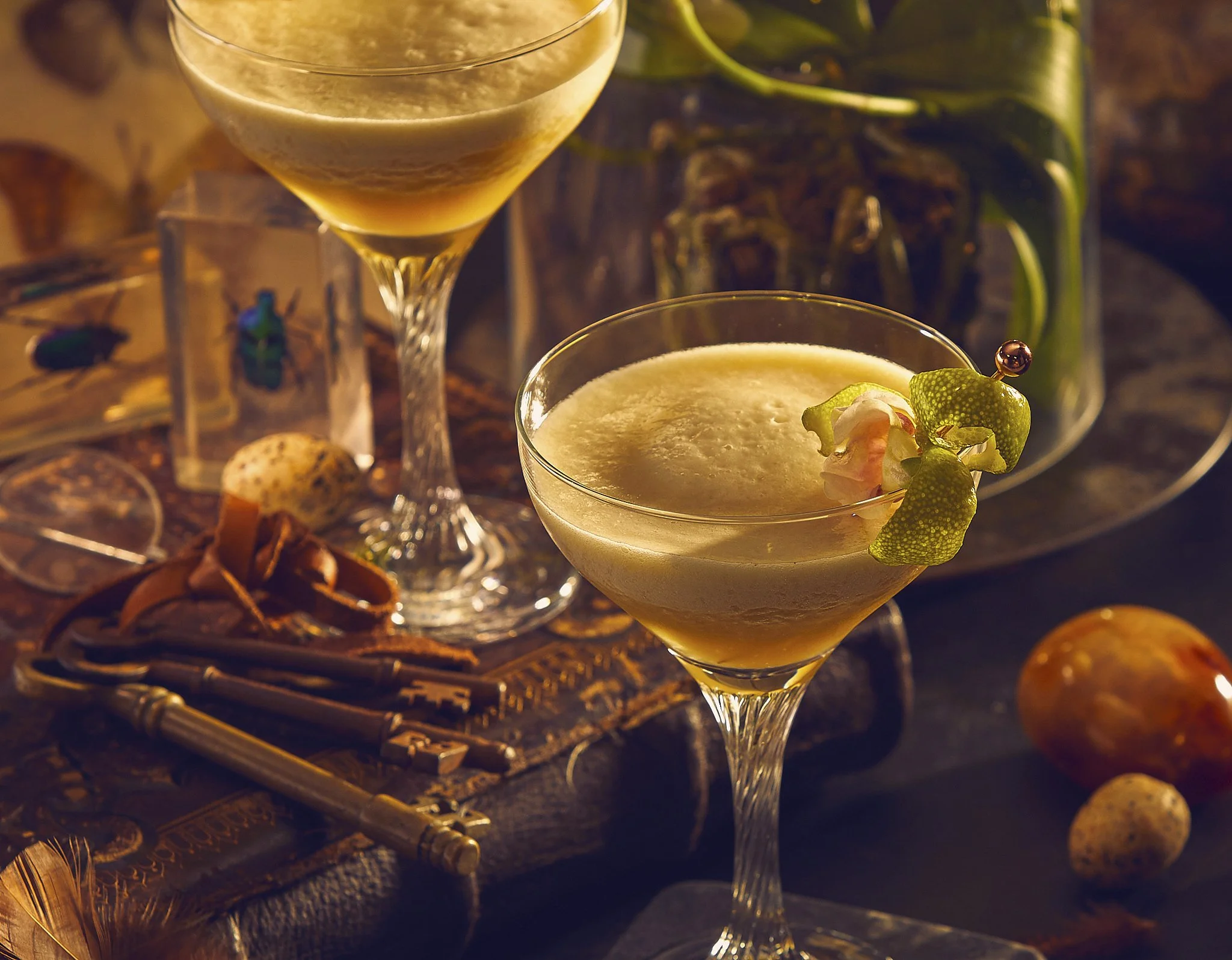 Two elegant cocktail glasses filled with a creamy, frothy beverage, garnished with a small flower and a green peel, set on a rustic wooden surface with decorative objects and plants in the background.