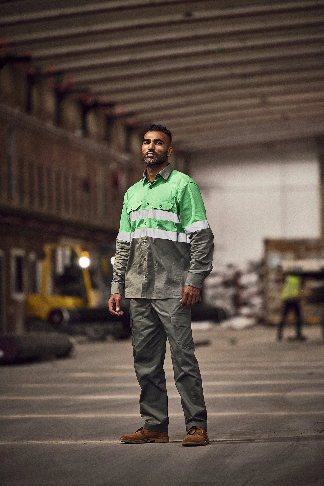 A man with a beard and short hair wearing a green and gray high-visibility work shirt and gray pants, standing in a warehouse or industrial setting. The background features storage racks and machinery, and the lighting is dim with a focus on the man.