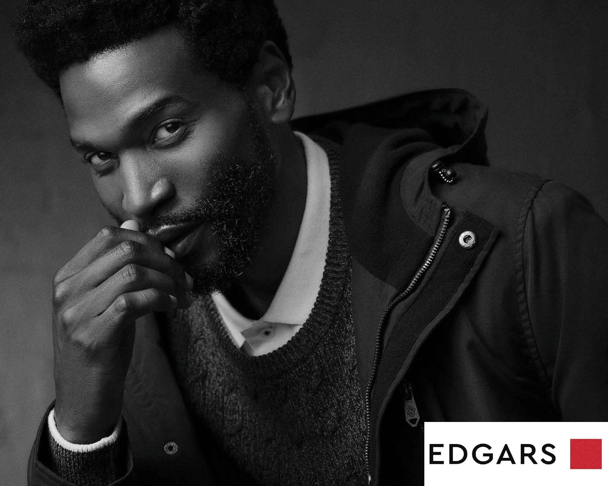 A black and white portrait of a young man with dark skin, curly hair, and a beard. He is looking directly at the camera with his hand near his mouth, and he is wearing a casual jacket over a shirt. The bottom right corner contains the logo for Edgar'