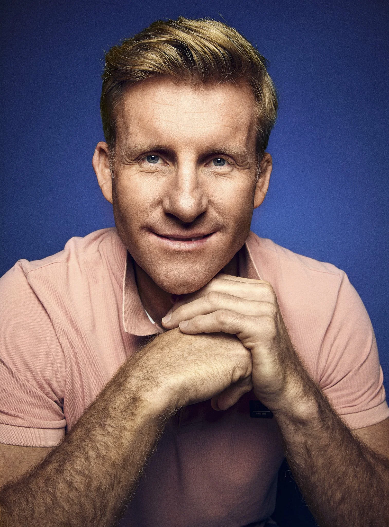 Close-up portrait of a AJ Venter with blonde hair and blue eyes, resting his chin on his hands, wearing a light pink shirt, against a dark blue background.