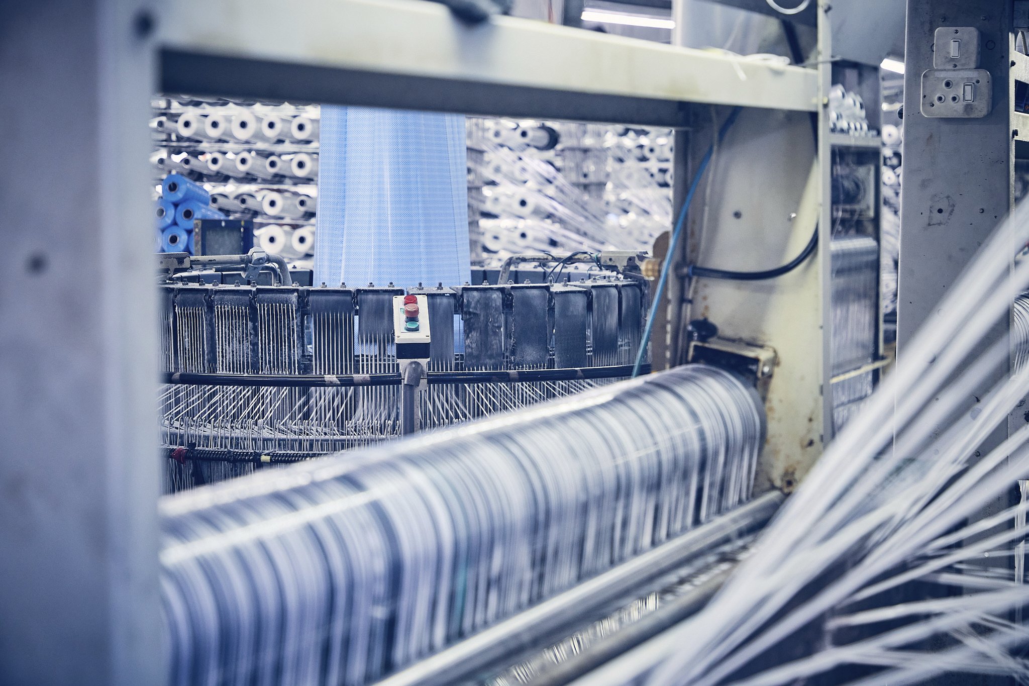 Close-up of a textile manufacturing machine with spools of thread in the background, some parts of the machine are visible including control buttons and wiring.