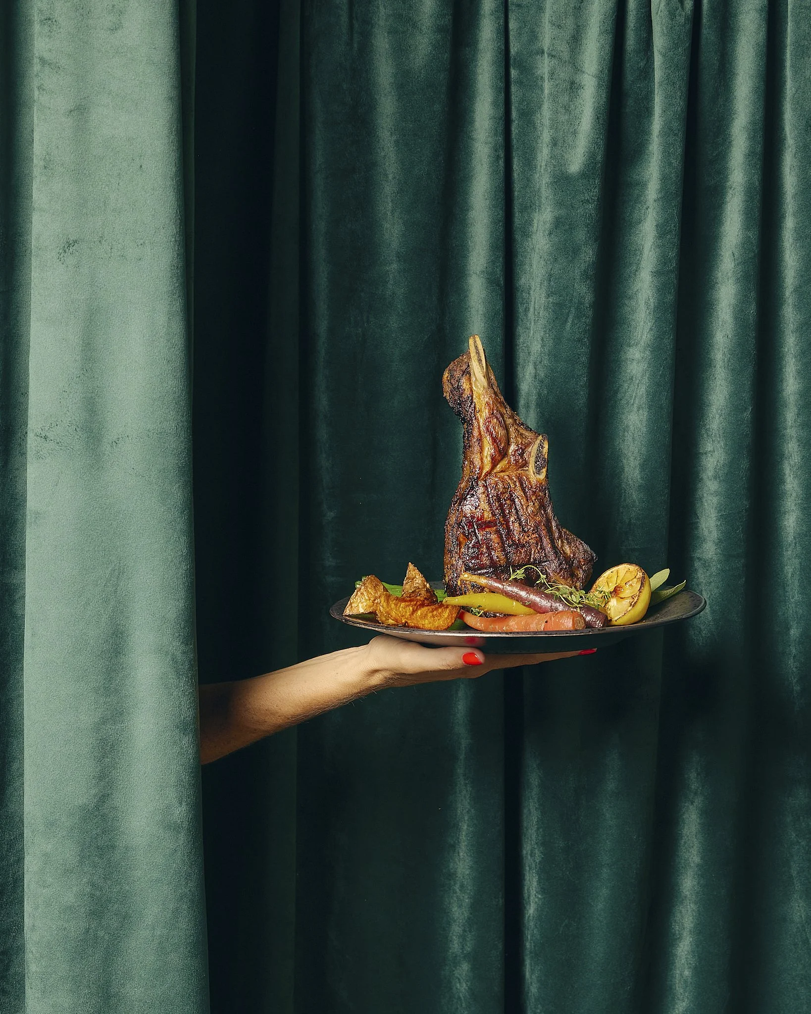 A person with red nail polish holding a plate of roasted meat and vegetables in front of green curtains.