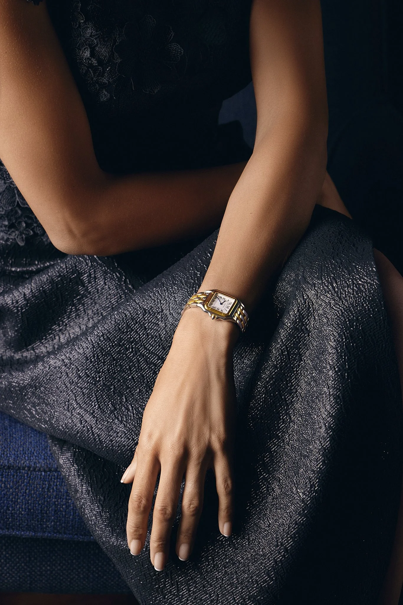 Close-up of a woman's arm and hand resting on her lap, wearing a gold Cartier wristwatch, black lace dress, and textured metallic fabric.