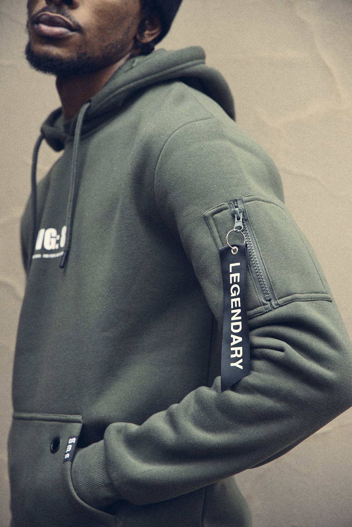 Close-up of a person wearing an olive green hoodie with a small zippered pocket on the sleeve, which has a black label with white text reading 'LEGENDARY', against a plain background.