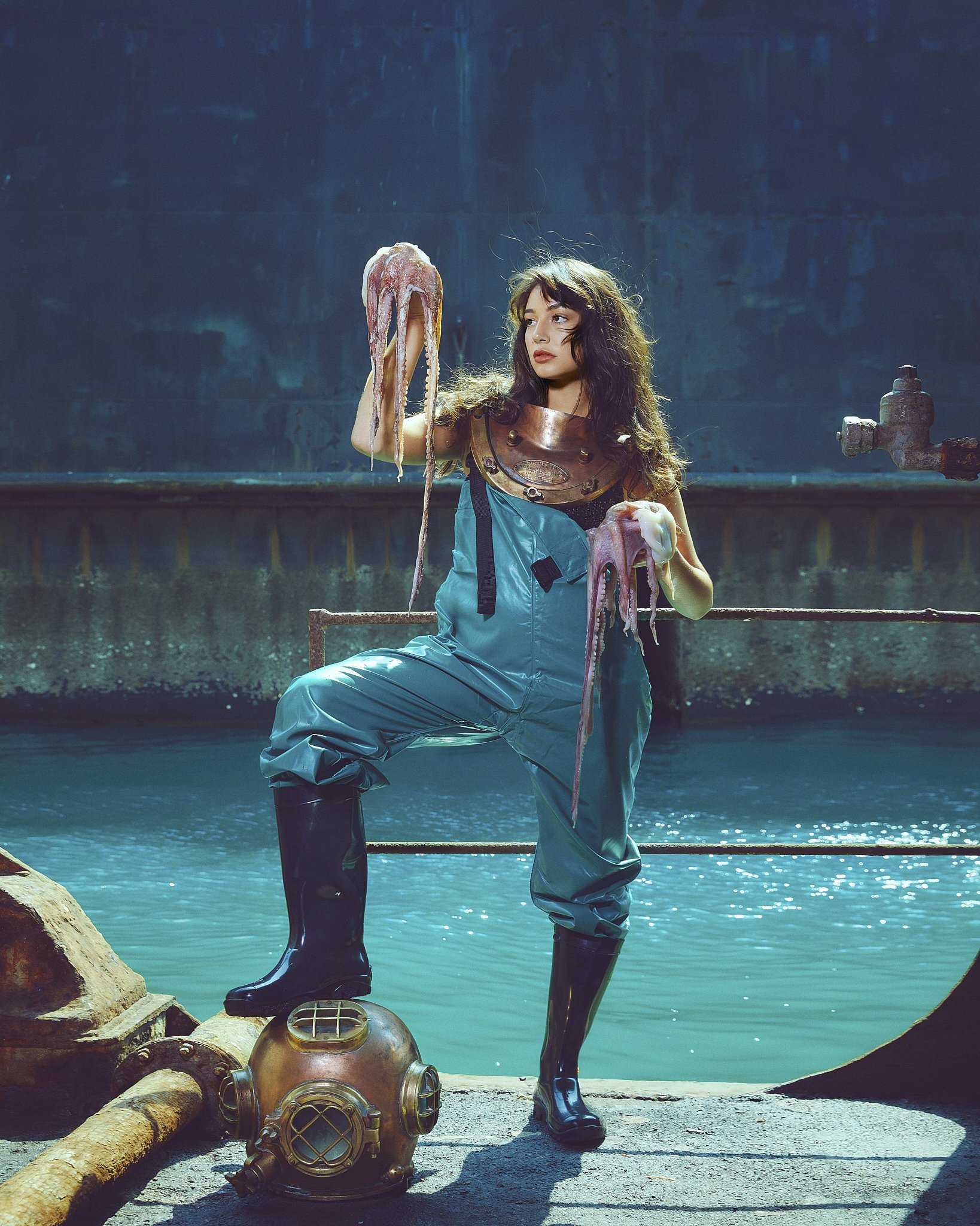 A woman dressed in a sci-fi or steampunk costume, wearing a metallic chest plate, blue pants, and boots, stands near water holding a model octopus with a similar steampunk design. There is an old-fashioned deep-sea diving suit helmet on the ground ne