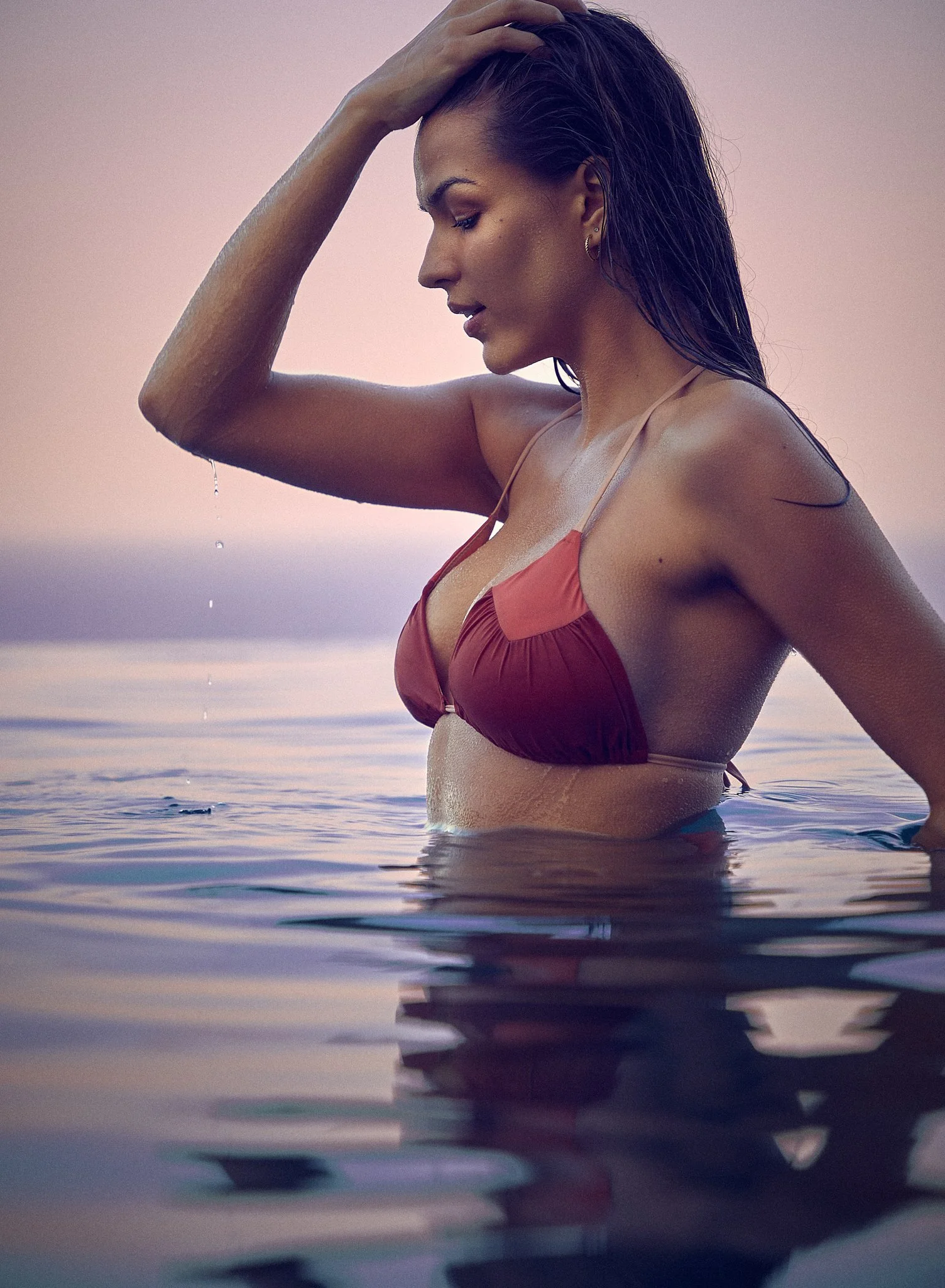 A woman in a red bikini is standing in shallow water during sunset, with her hand resting on her head and water dripping from her arm.