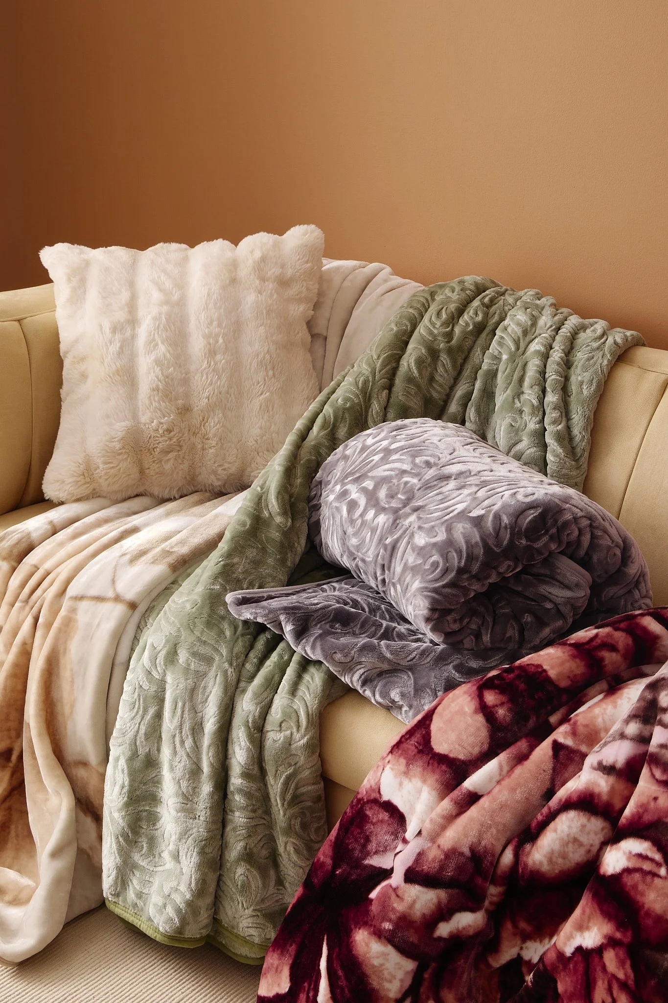 Various textured Sheet Street blankets and pillows draped over a beige sofa against a warm-colored wall.