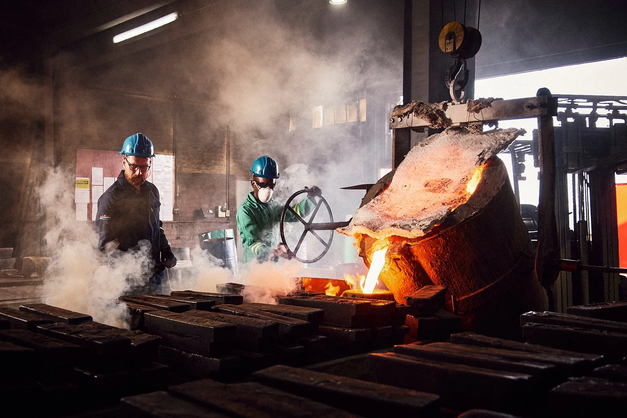 Two workers in protective helmets and gloves operate a furnace pouring molten metal into molds in a factory setting with smoke and bright orange glow.