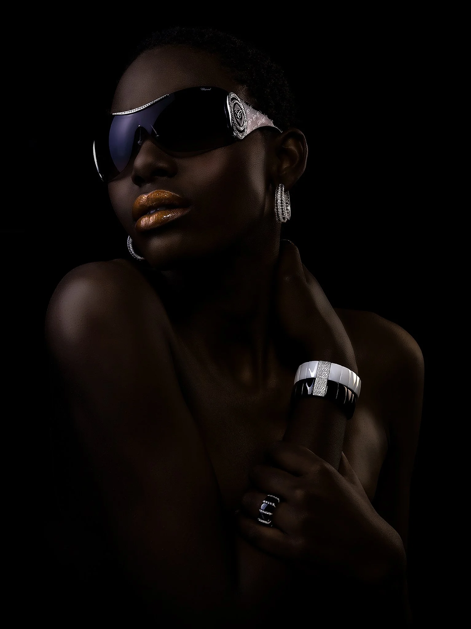 A close-up portrait of a woman with dark skin wearing large dark sunglasses and gold lipstick, accessorized with silver jewelry, set against a black background.