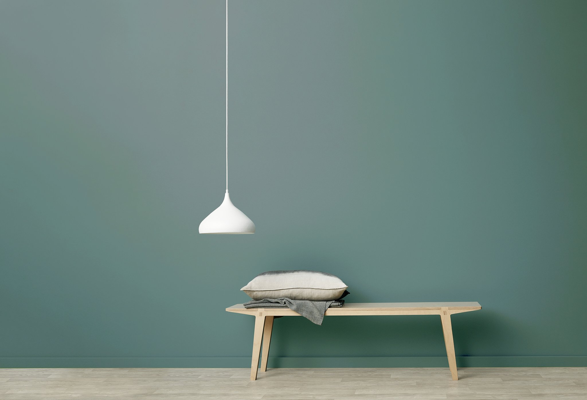 A minimalist interior with a teardrop-shaped white pendant light hanging above a wooden table. On the table, there is a stack of folded fabrics and a cushion. The background is a plain teal wall with a simple light-colored wooden floor.