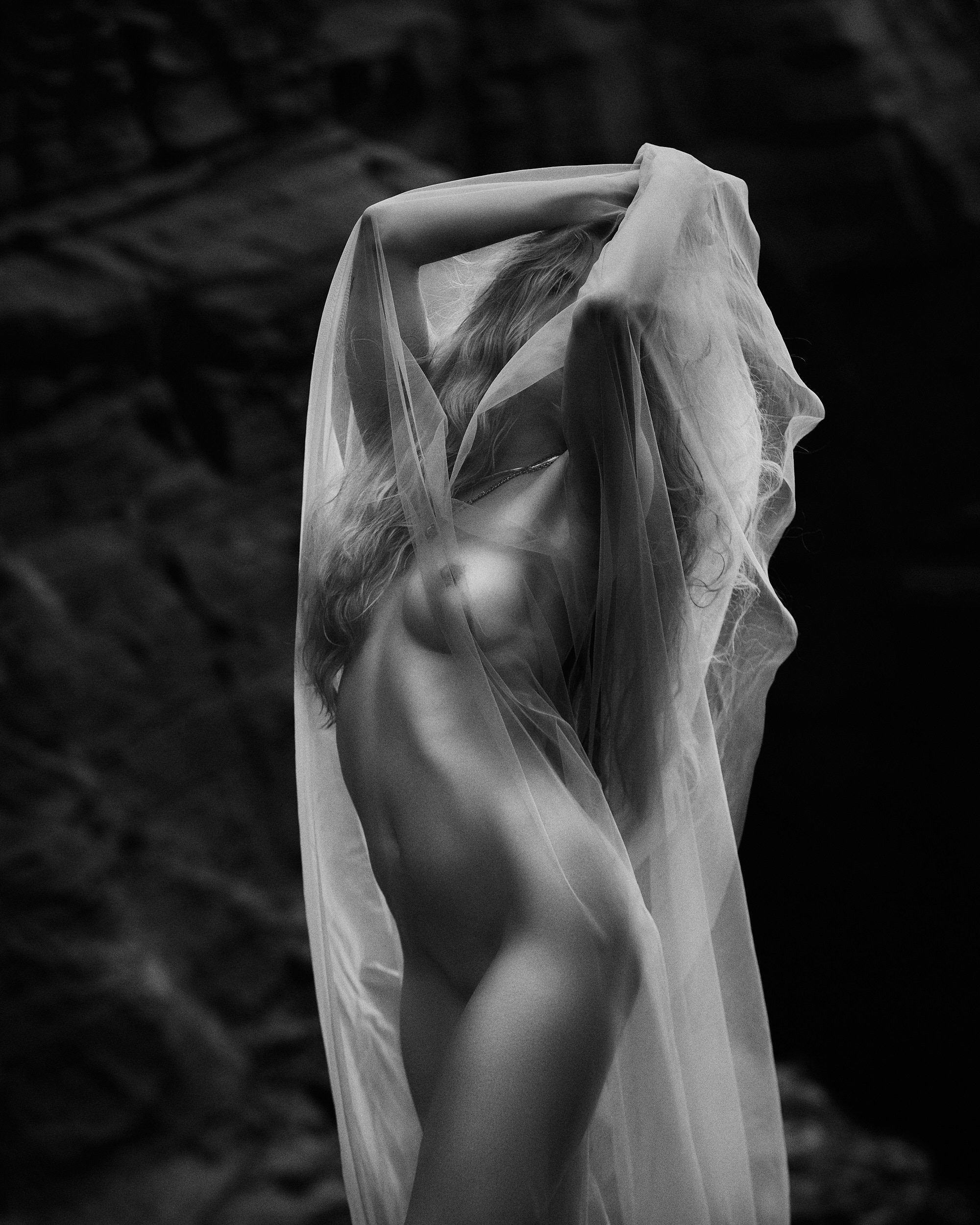 A nude woman draped in a sheer fabric, with her arm raised, partially covering her face, in a black and white photo against a dark, blurred background.