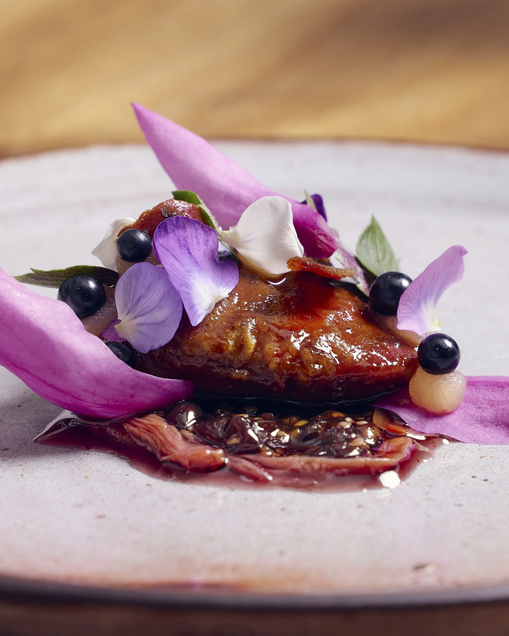 Close-up of a gourmet dish featuring a piece of glazed meat garnished with purple and white flower petals, small dark berries, and edible leaves, presented on a white plate.