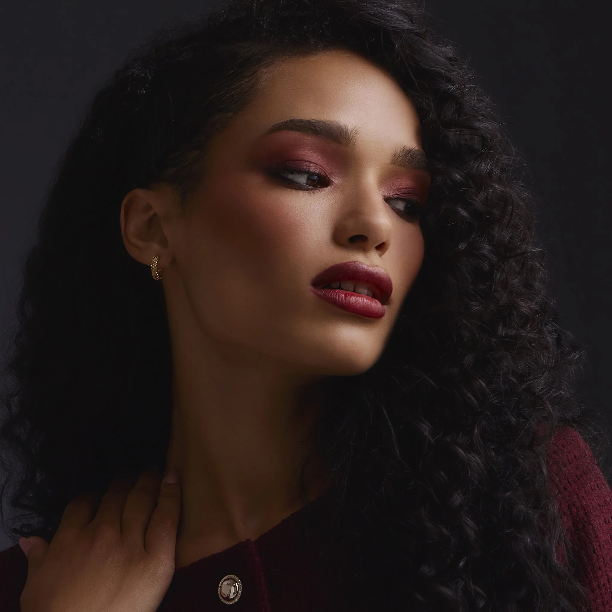 A woman with dark, curly hair and pink eye makeup, wearing a maroon top, a gold earring, and a ring with a decorative band, is touching her neck with her hand.
