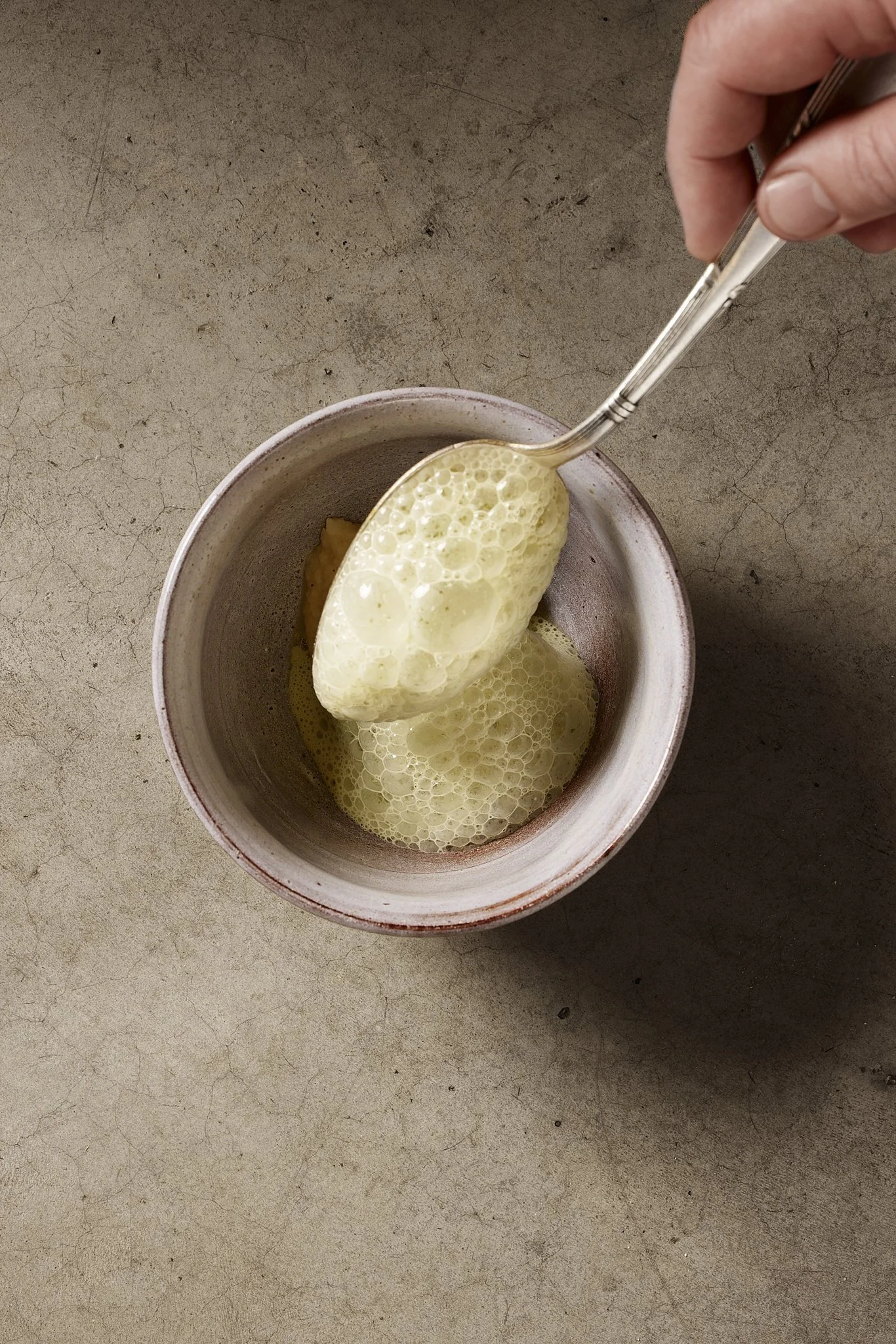 A hand using a silver spoon to scoop yellow foam from a mug on a textured beige surface.