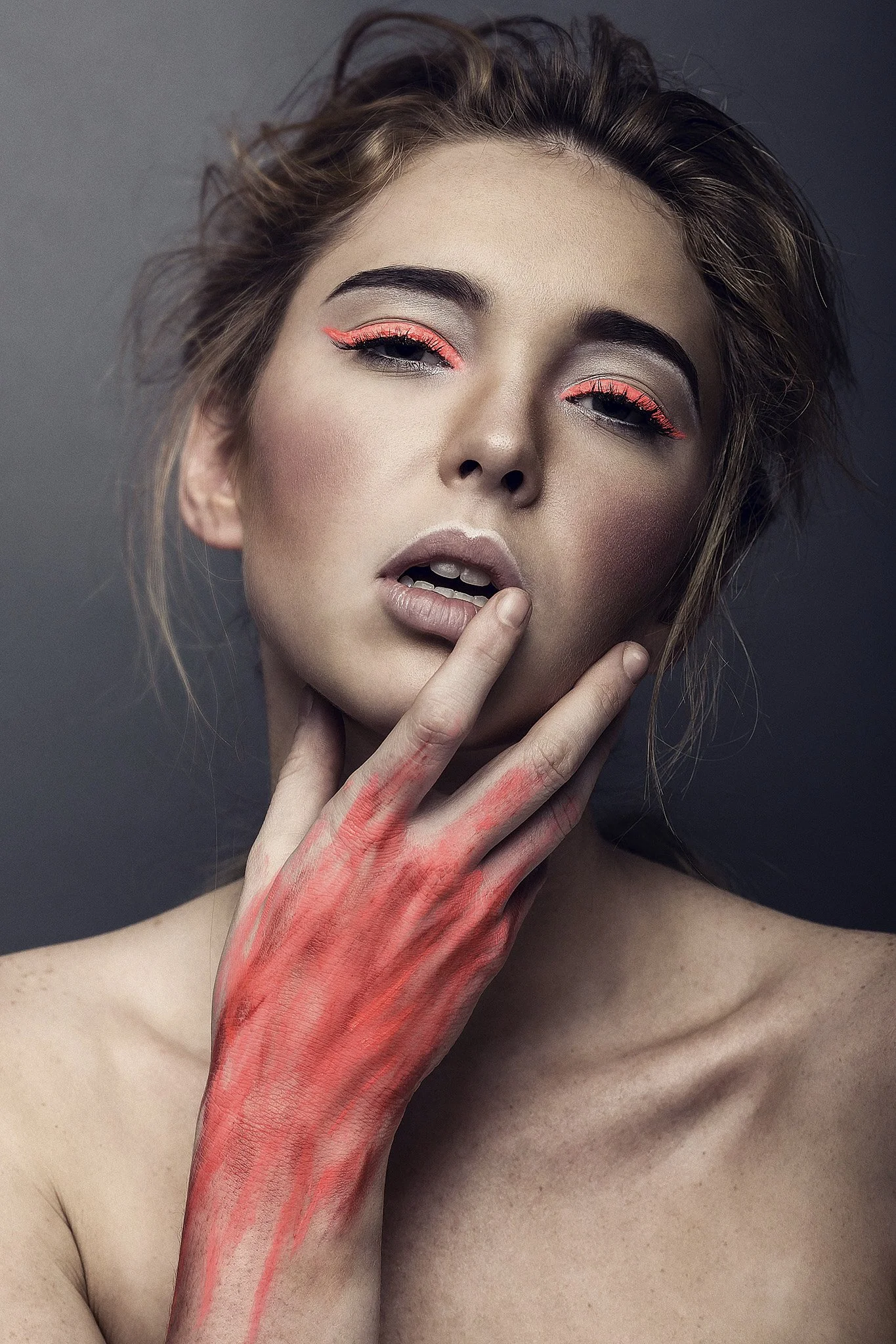 A close-up portrait of a woman with eye makeup and lipstick, holding a red painted hand near her face, with a neutral background.