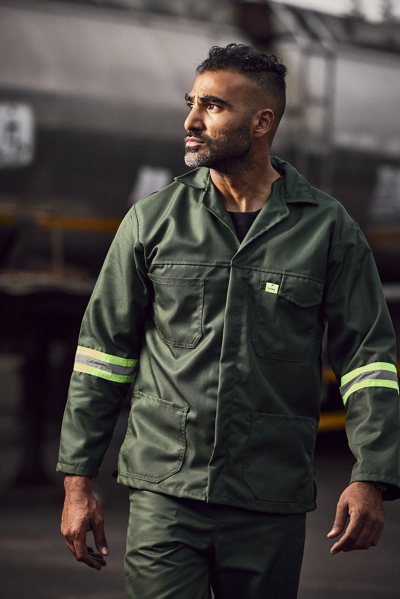 A man in a green work uniform with reflective stripes on the sleeves standing in a warehouse or industrial setting.