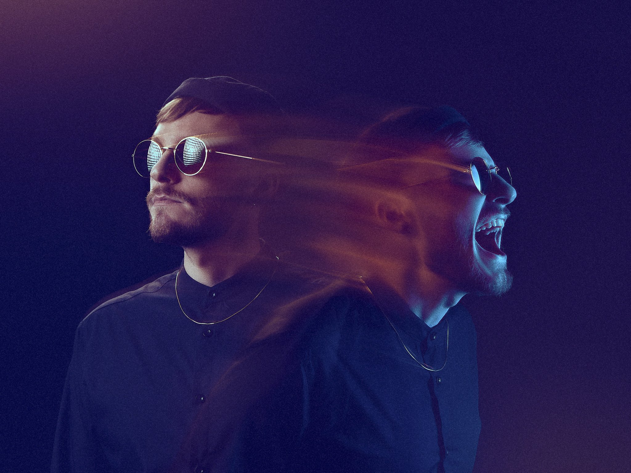 One man long exposure wearing sunglasses and black shirts, person on the left has a beard, person on the right has a beard and is smiling with mouth open, both facing opposite directions, with colorful light streaks in the background.