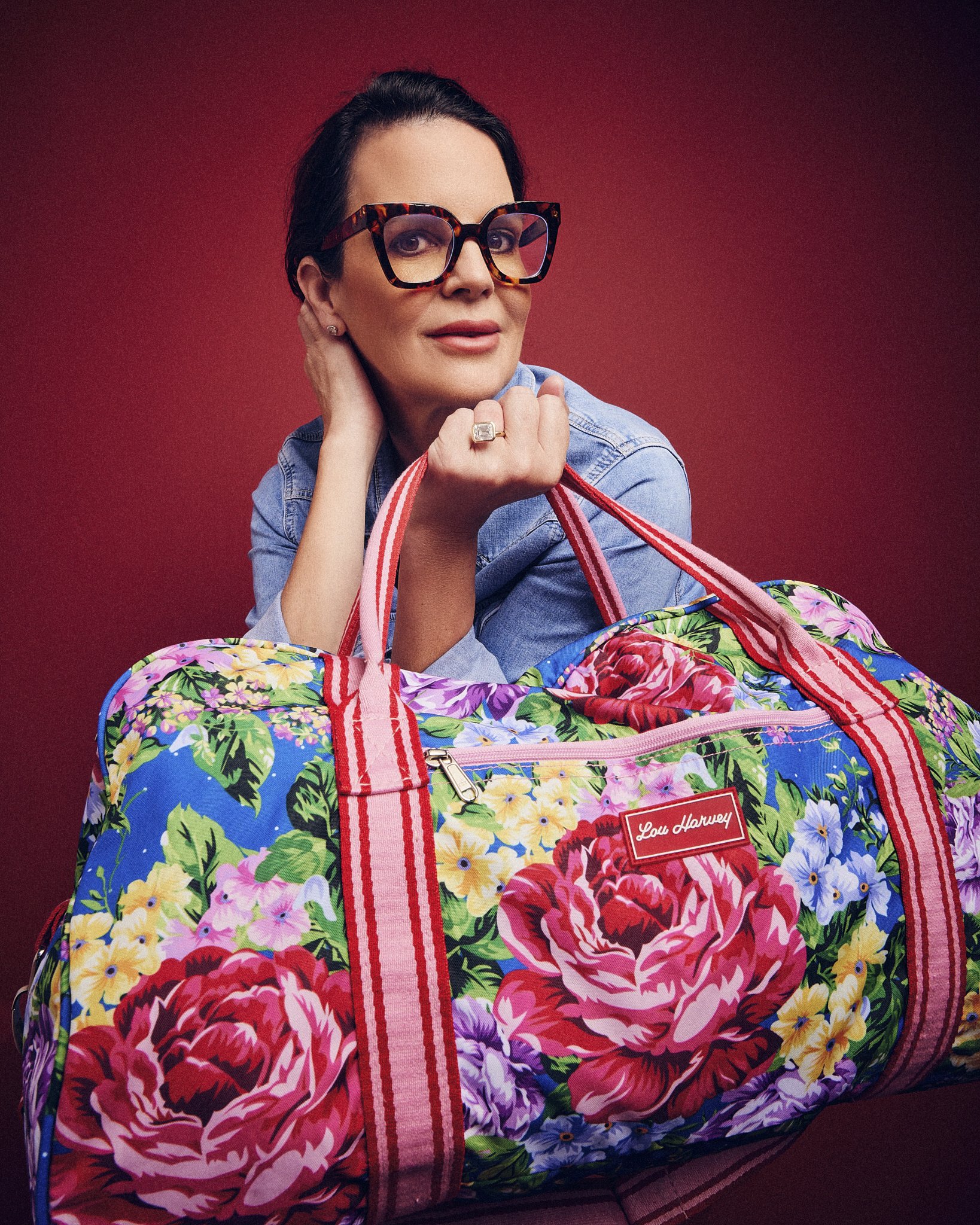 A woman with short dark hair wearing large tortoiseshell glasses, a denim jacket, and earrings, resting her chin on her hand, looking at the camera, against a red background. She has a ring on her finger. In front of her, there is a colorful floral-p