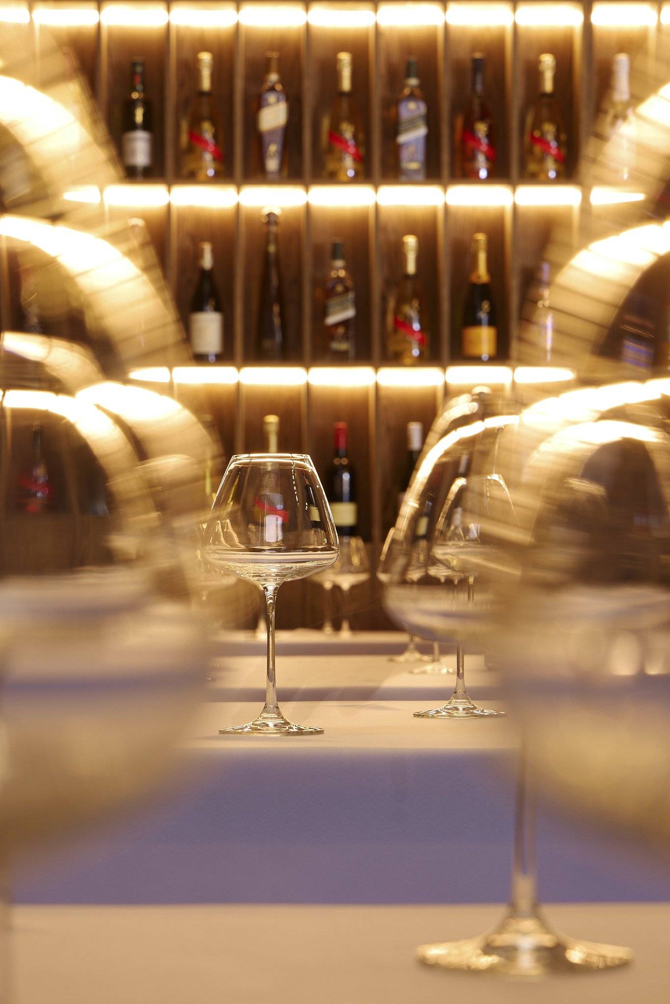 A row of empty stemware glasses on a table with blurred wine bottles in a wine bar setting in the background, illuminated by warm lighting.