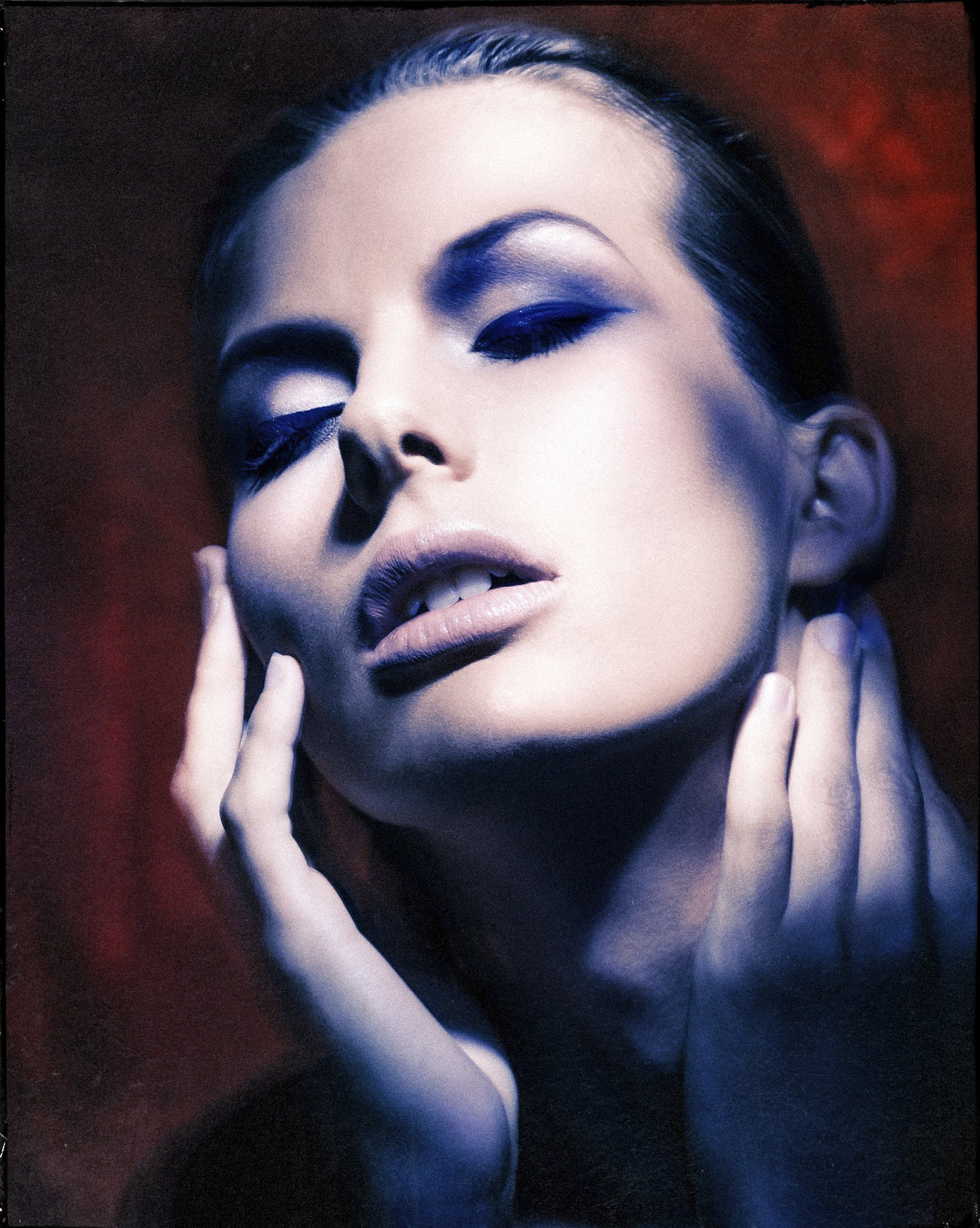 Close-up of a woman with closed eyes, dark hair, and makeup, with hands gently touching her face against a dark, red and black blurred background.
