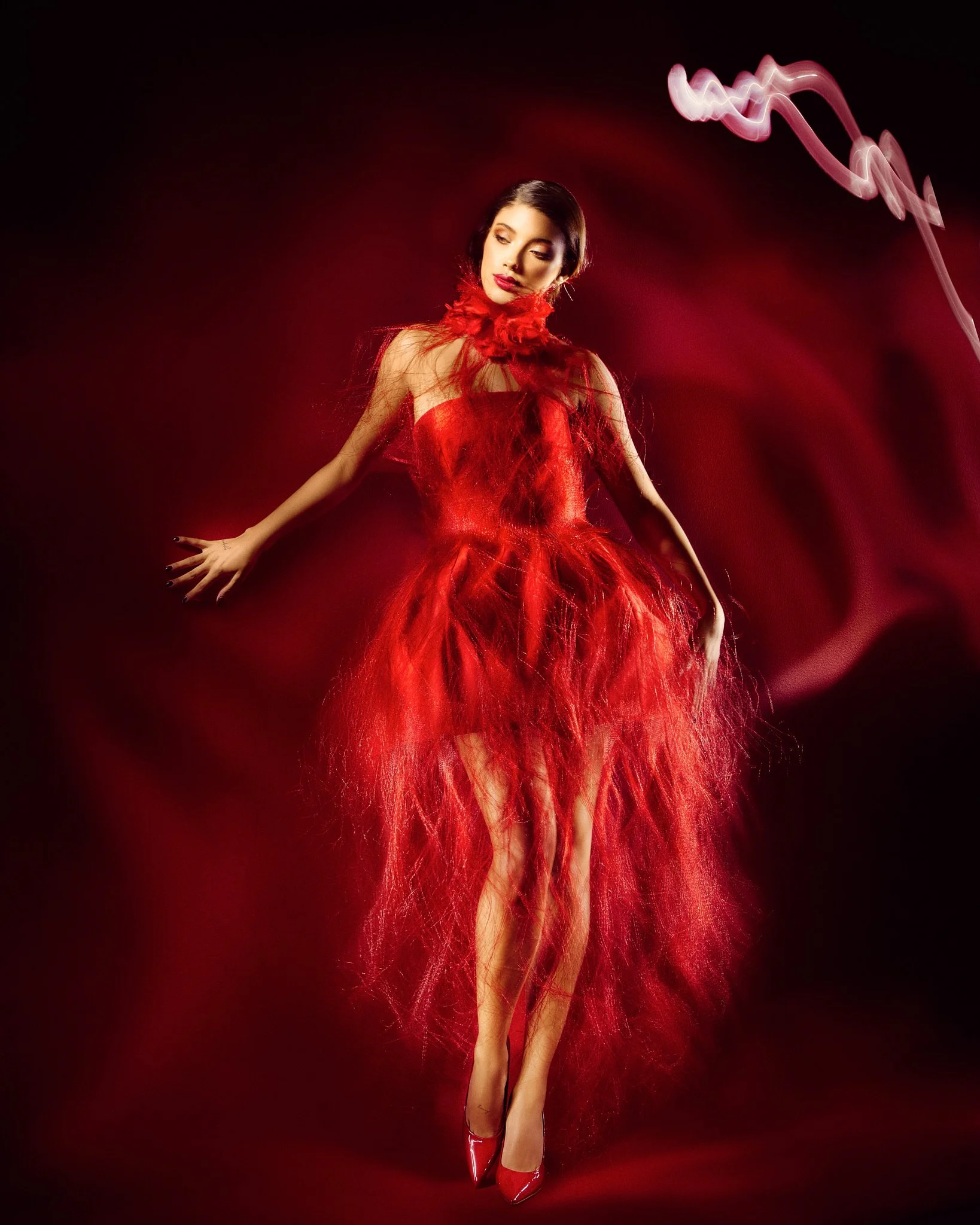 A woman with short dark hair poses against a black background with red lighting and magenta light streaks. She wears a red dress made of feathers and red high heels, and her arms are extended outward with her hands relaxed. painting with light