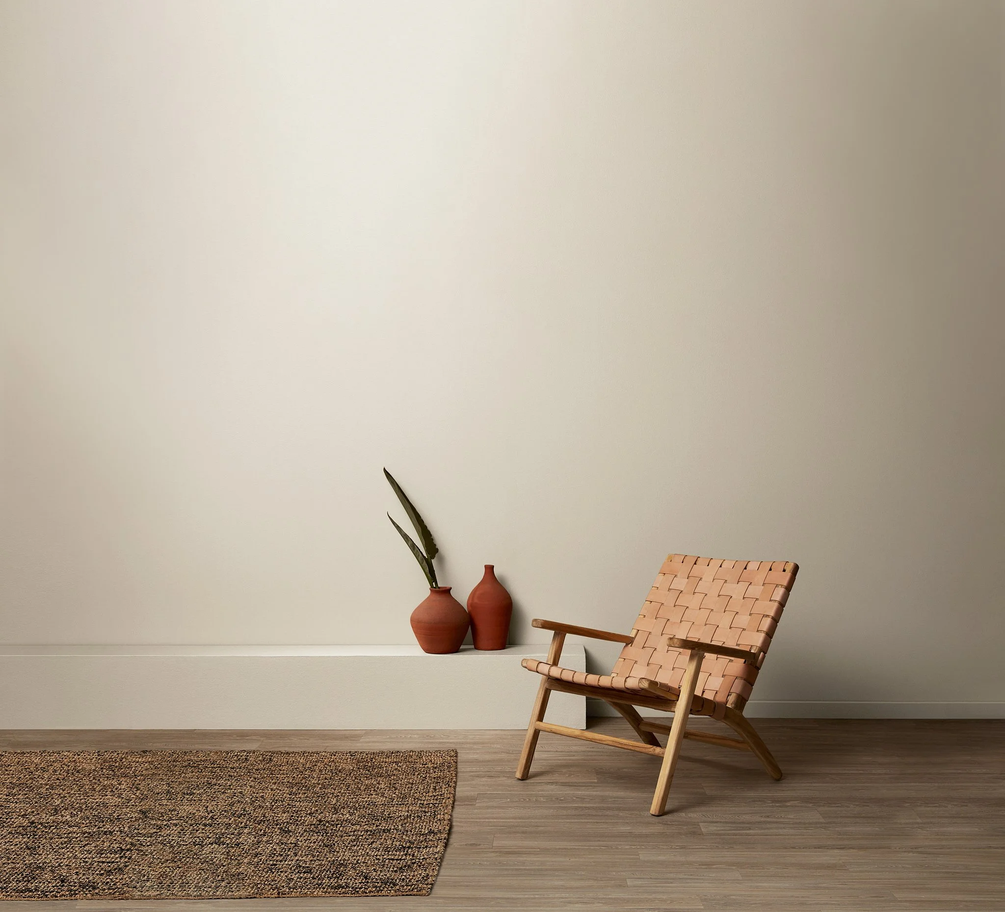 A minimalist interior of a room with a beige wall, a low white shelf with three terracotta vases, a wicker chair with a wooden frame, and a patterned rug on hardwood flooring.