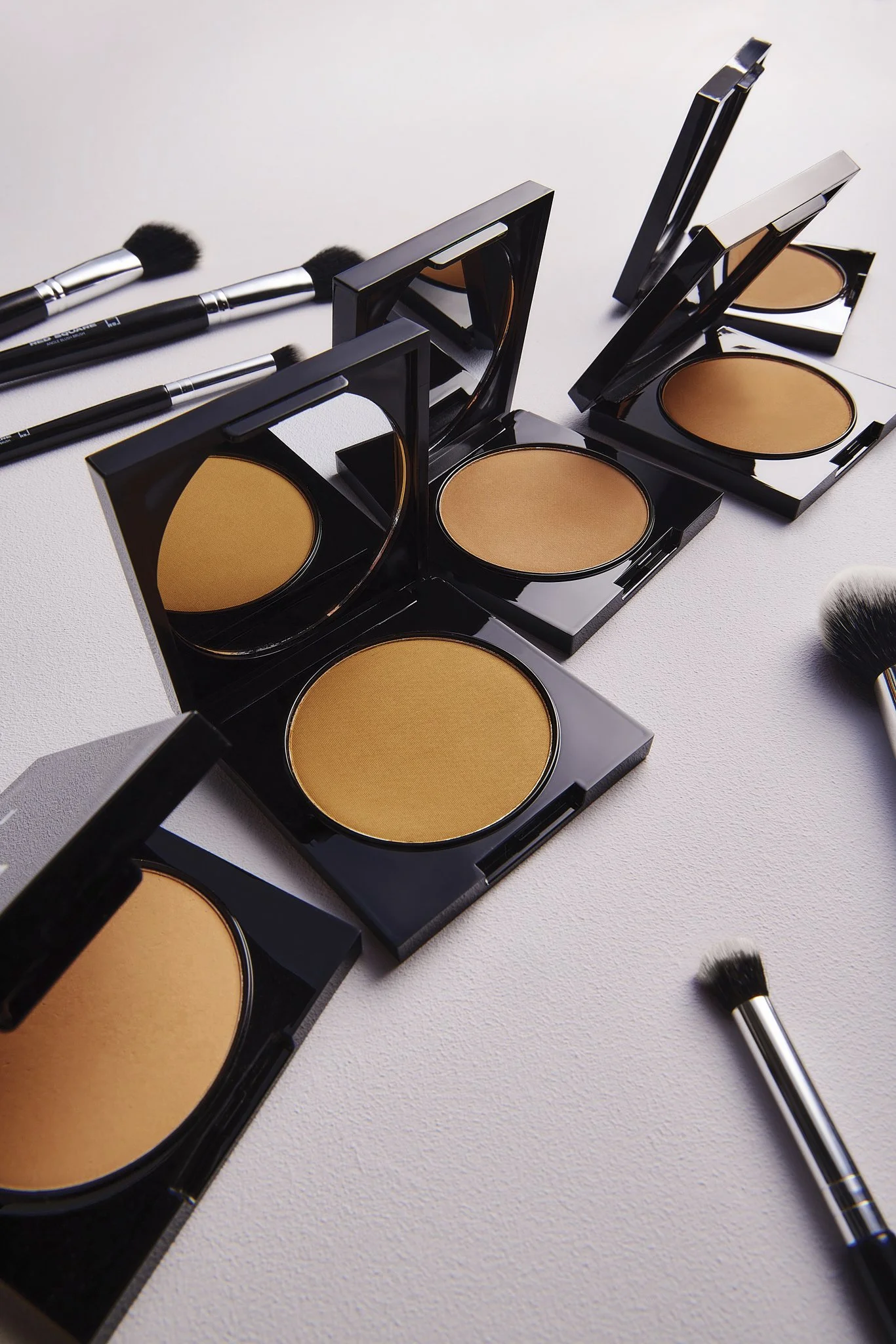 Beauty setup with three compact powders in black cases and several makeup brushes on a light-colored surface.