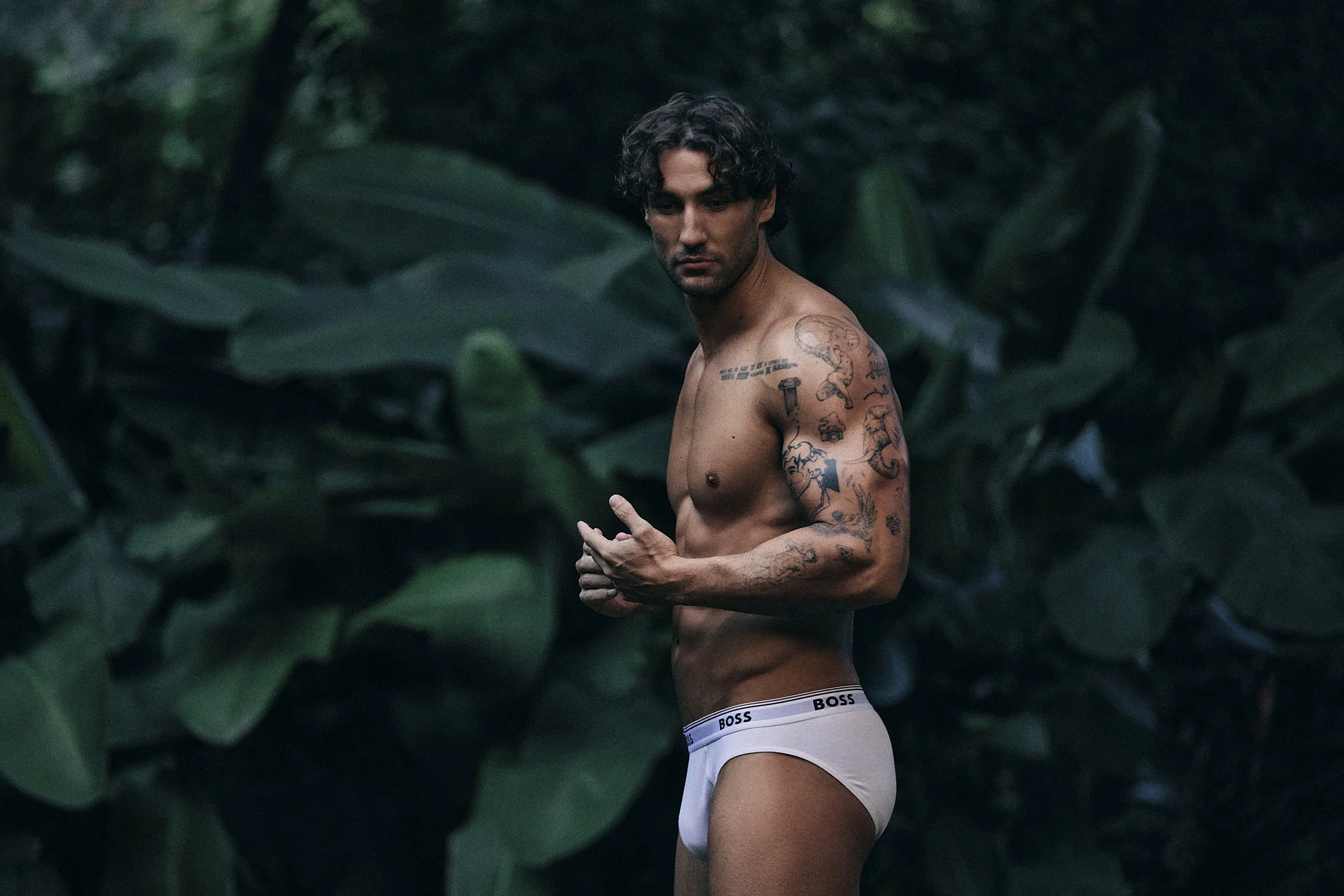 A shirtless man with curly dark hair and tattoos on his arms and shoulder posing among green foliage, wearing white underwear with the brand 'BOSS' visible on the waistband.