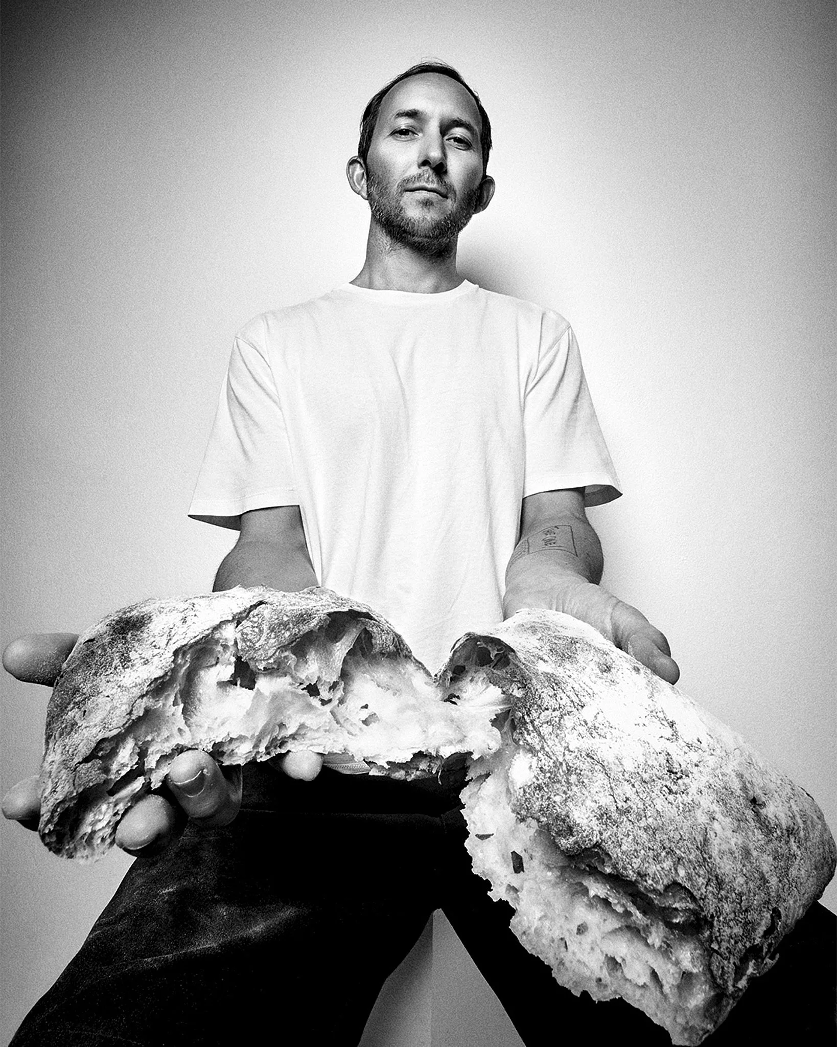 Wide angle portrait black and white photograph of of the baker with a beard and short hair, holding a large, broken open bread sourdough loaf, with a serious or neutral expression, against a plain background.