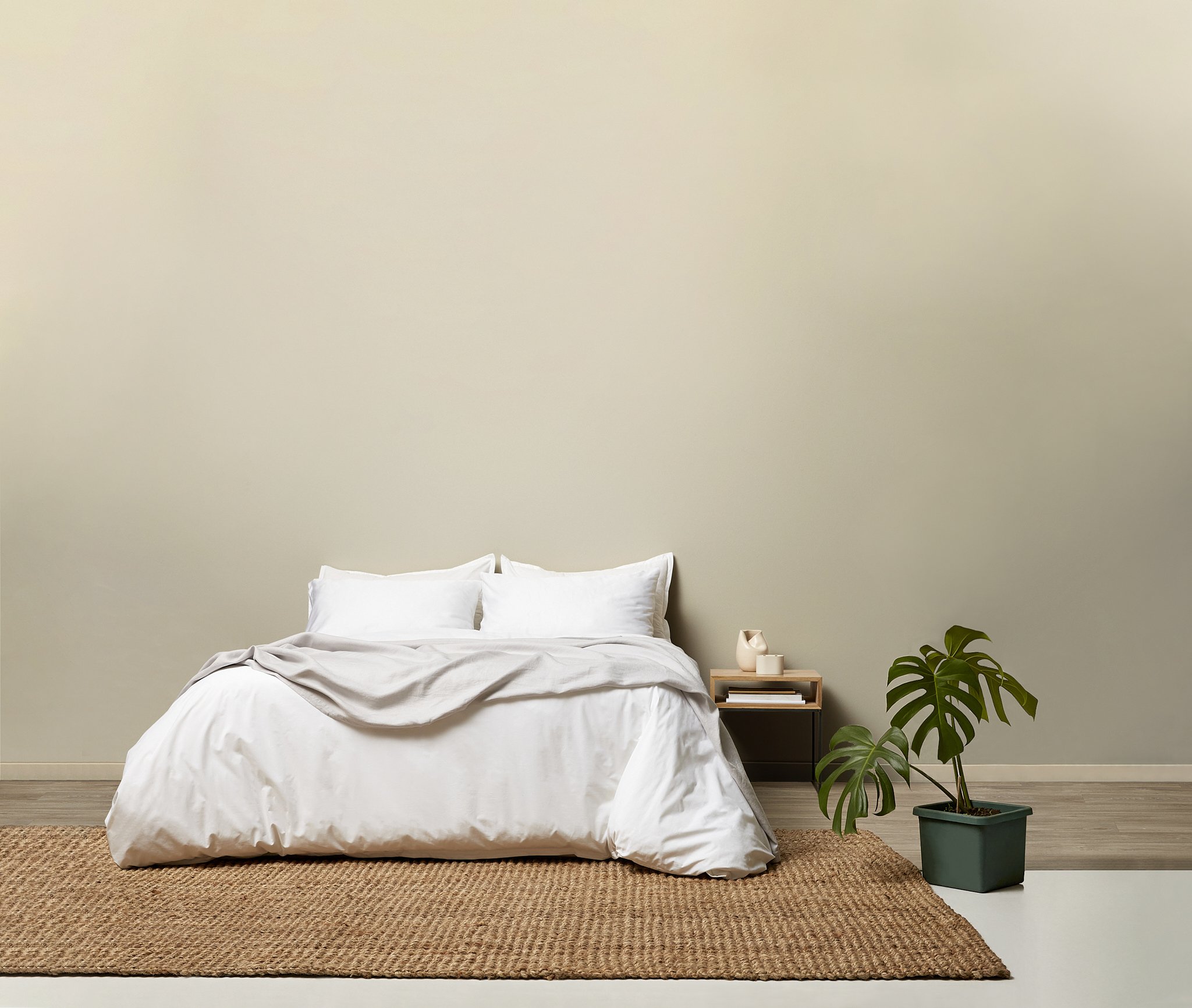 A minimalistic bedroom with a bed dressed in white linens, a wooden side table with a small decorative object and books, a large green potted plant, a beige textured rug, and a plain light-colored wall.